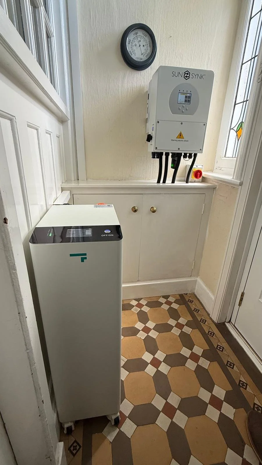 A small electric vehicle charger in a hallway with patterned tile flooring, a cabinet, a wall-mounted solar inverter, a clock, and an electrical switch.
