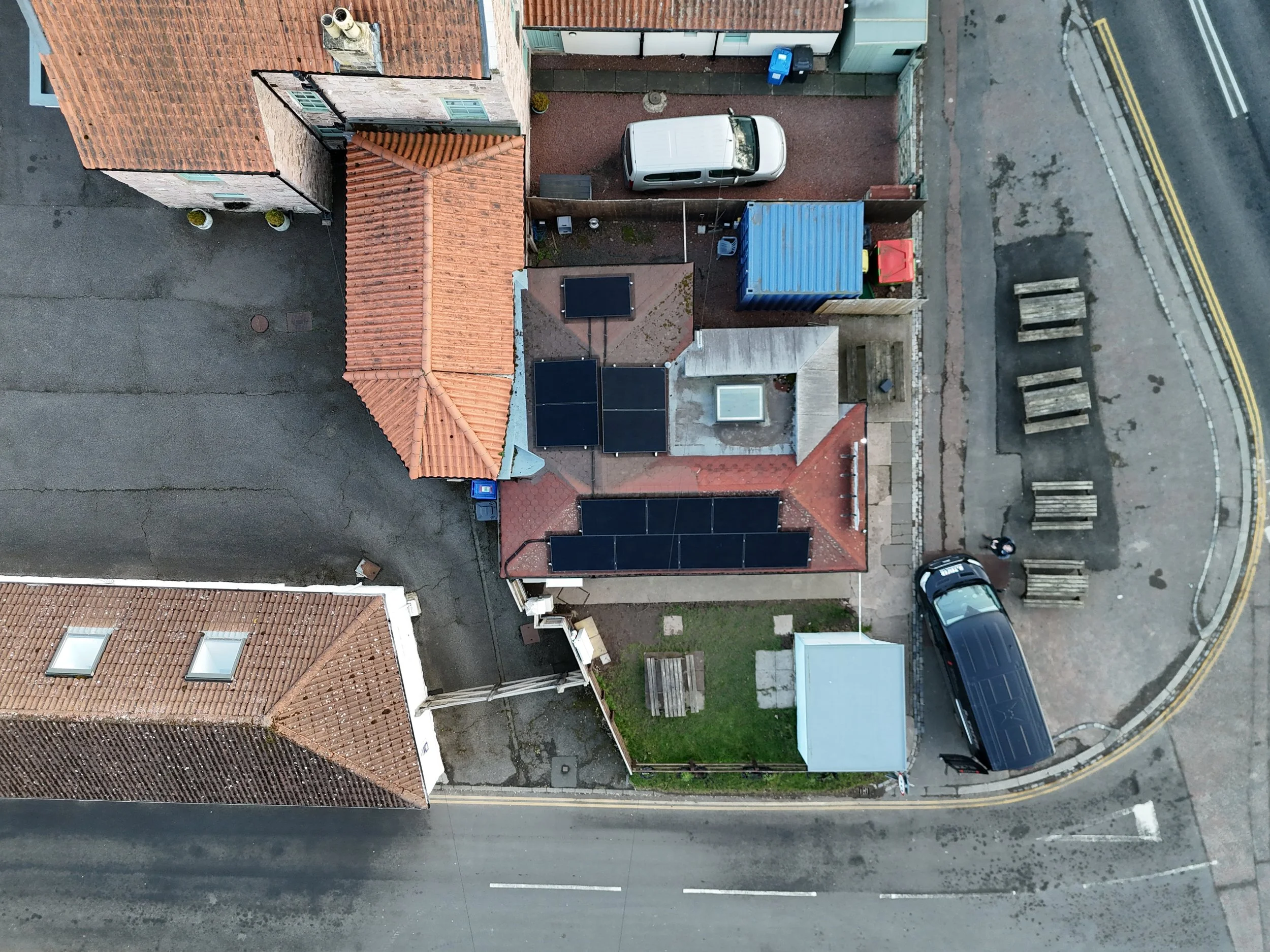 An aerial view of residential houses with red-tiled roofs along a street, showing a driveway with a white van, solar panels on the roof, and parked cars.