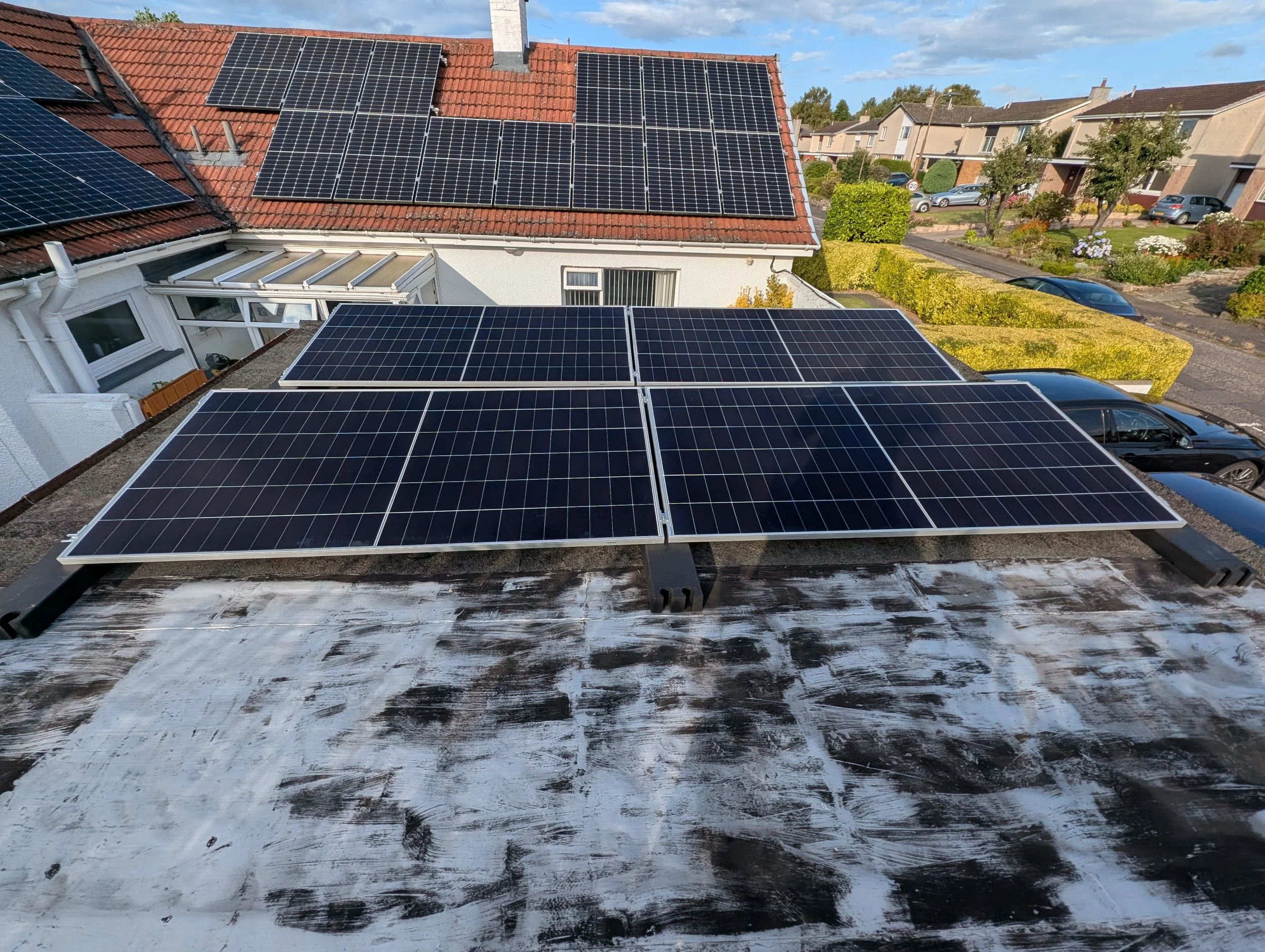 Roof with multiple solar panels in a residential area, with a view of neighboring houses, trees, and parked cars under a partly cloudy sky.