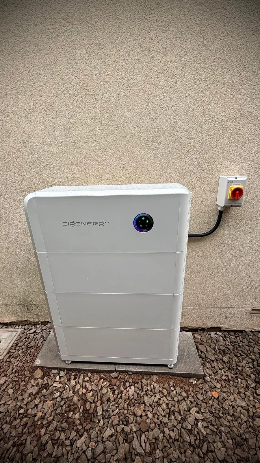 A white solar energy storage system with a circular digital display on the front, placed on a concrete pad outside a building with beige textured wall. It is connected to an electrical box on the wall with a black cable.