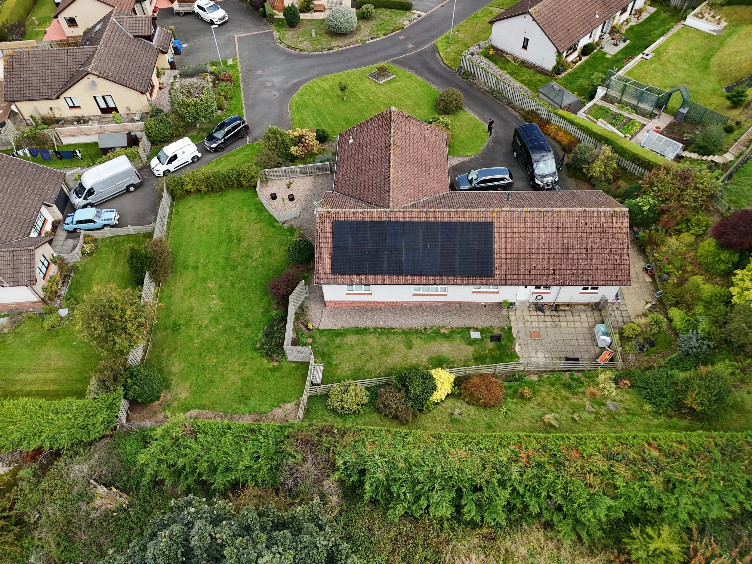 Aerial view of a suburban neighborhood showing houses, garages, cars, and well-maintained gardens with trees and bushes.