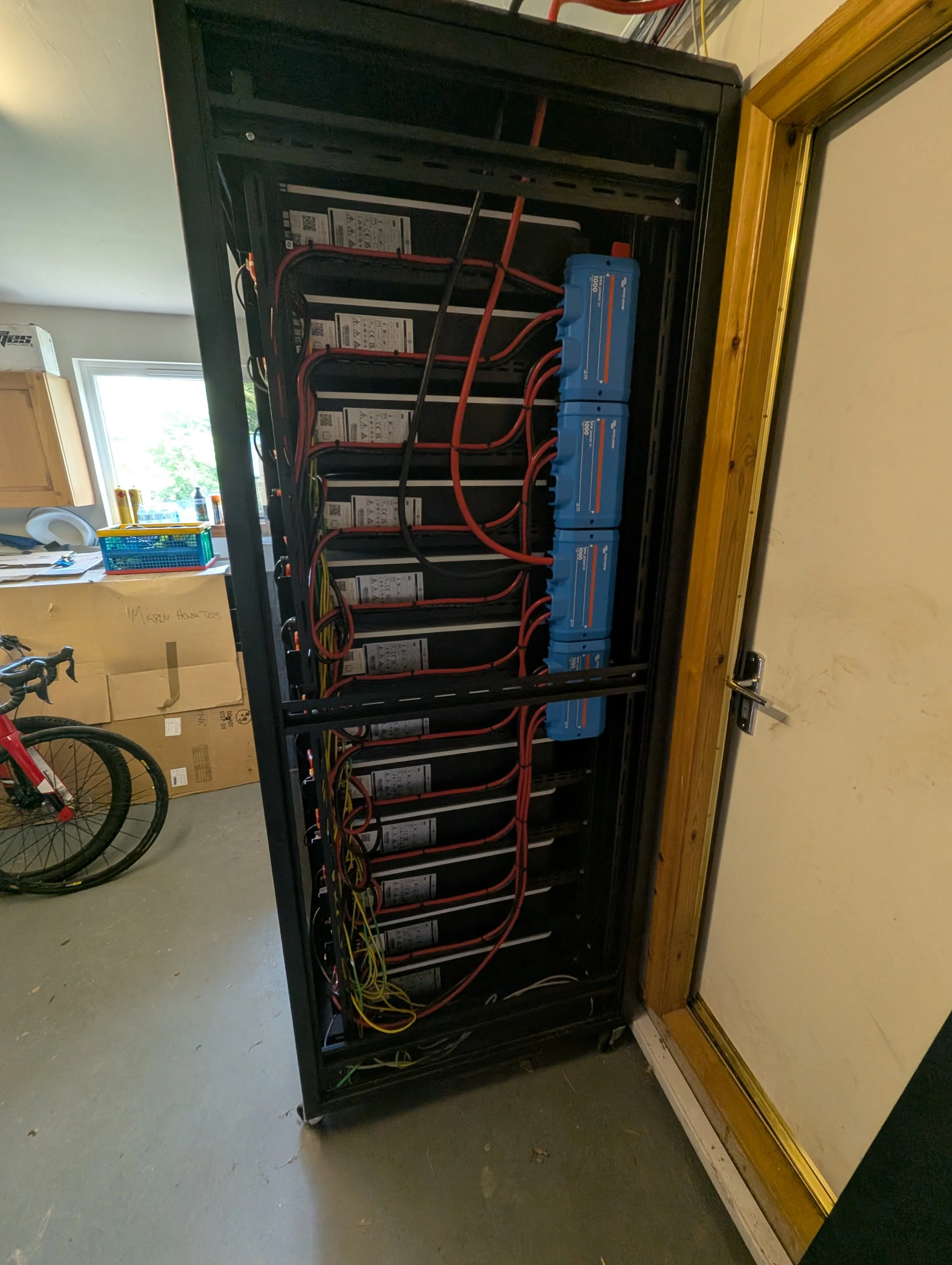 An open electrical cabinet containing batteries and wiring, set in a room with a window and cardboard boxes in the background.
