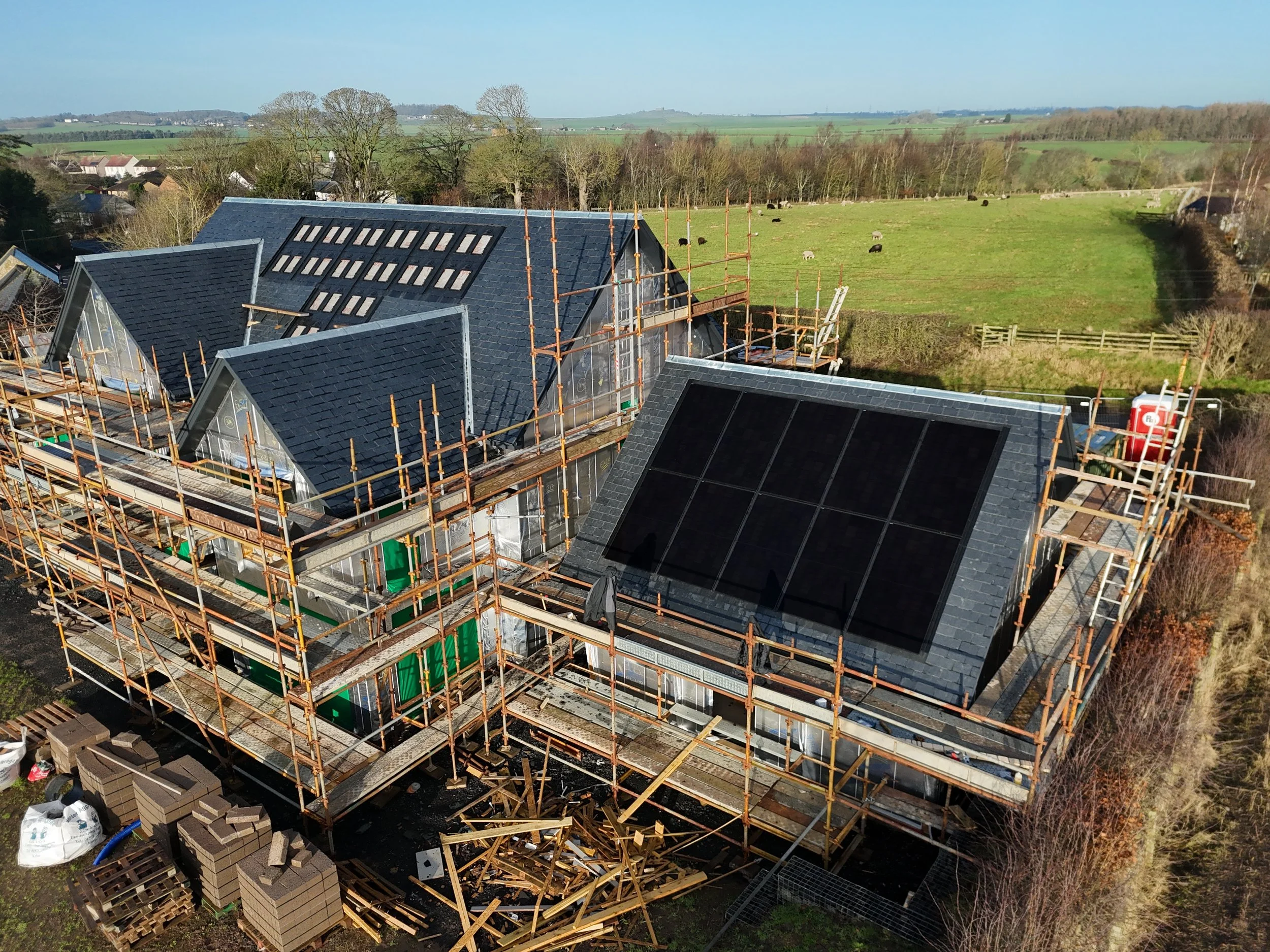 Aerial view of a house under construction with scaffolding, roofing in progress, and construction materials and bricks around the site, set in a rural landscape with fields and trees.