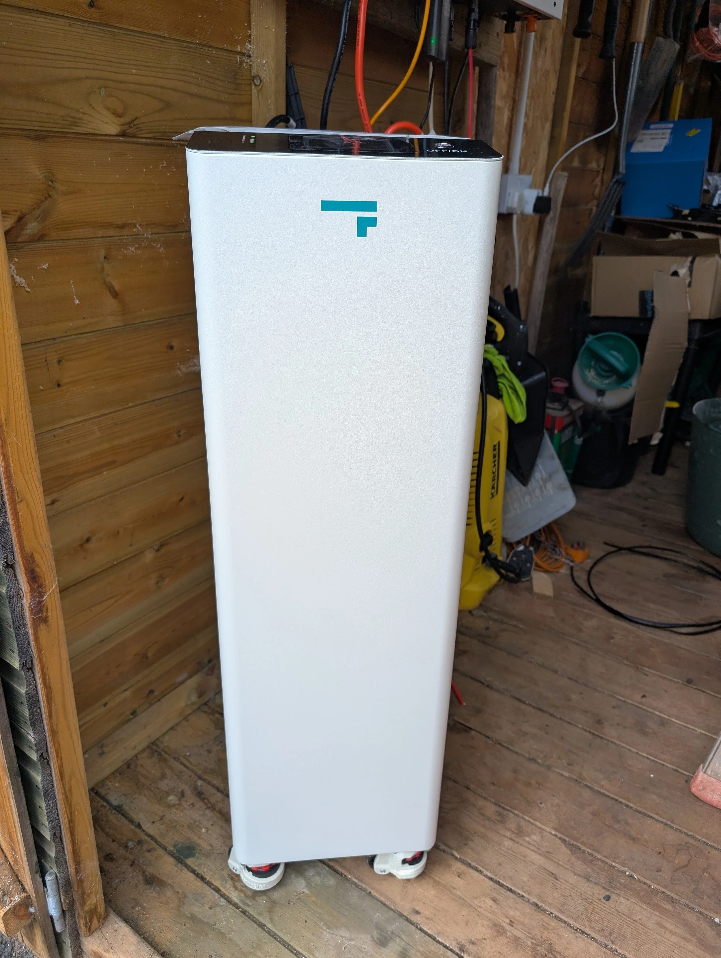 A white device with a black top and a blue logo on the front is situated on a wooden floor near a wooden wall. The device has four wheels, and there are various tools and cables in the background, including a yellow vacuum and other equipment.