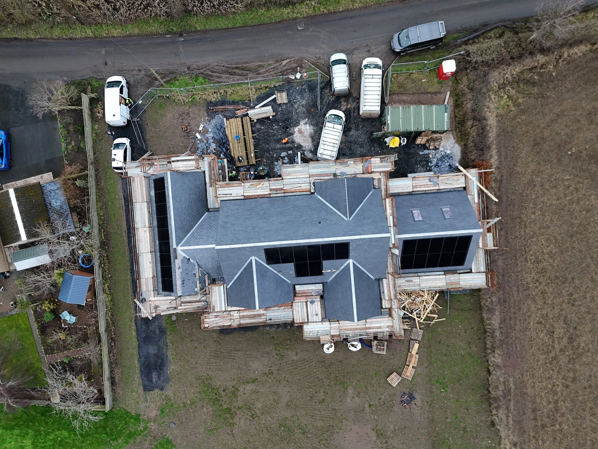 An aerial view of a house under construction with several construction vehicles and materials around, a partially paved driveway, and a dirt road in the background.