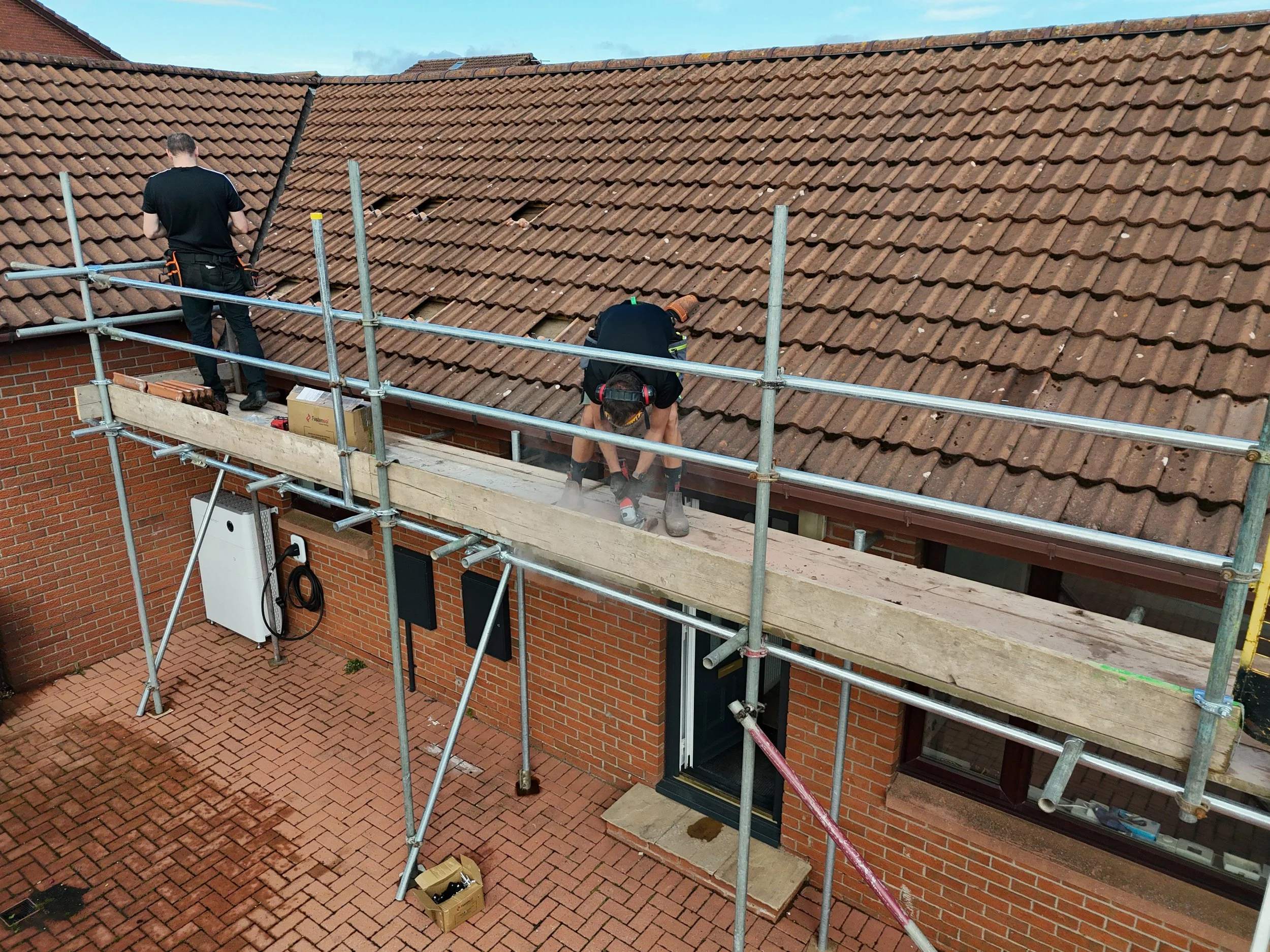 Two workers on scaffolding repairing a tiled roof of a brick building.