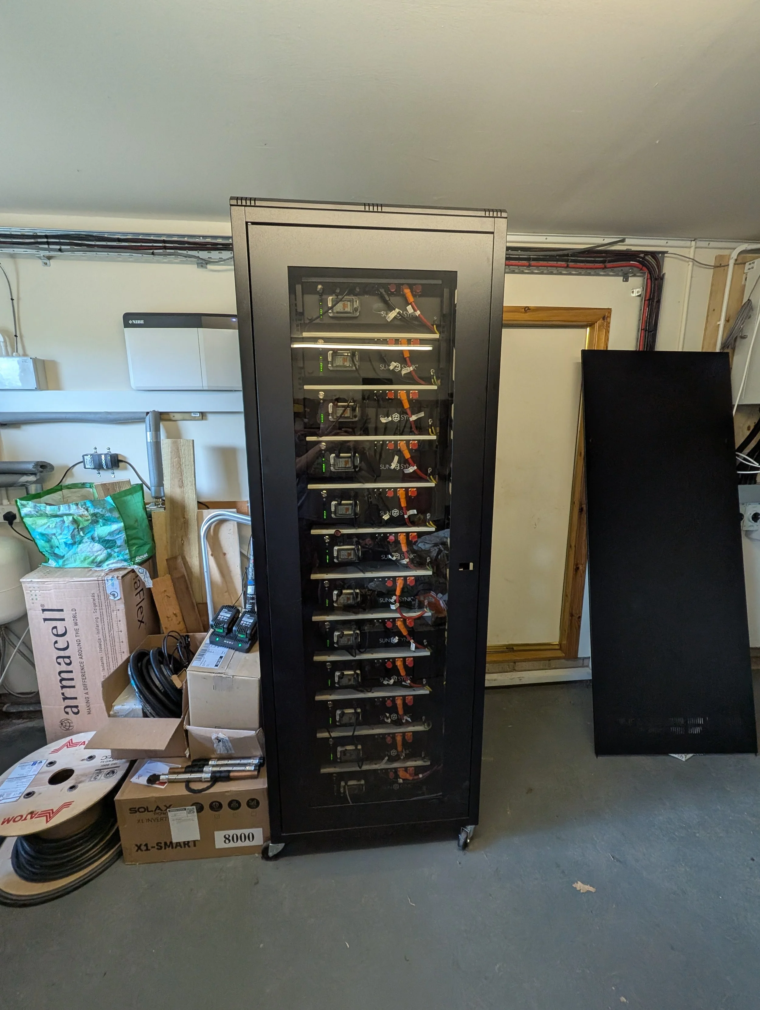 A black server rack with multiple shelves containing electronic components, situated in a room with various boxes, wires, and equipment around it.