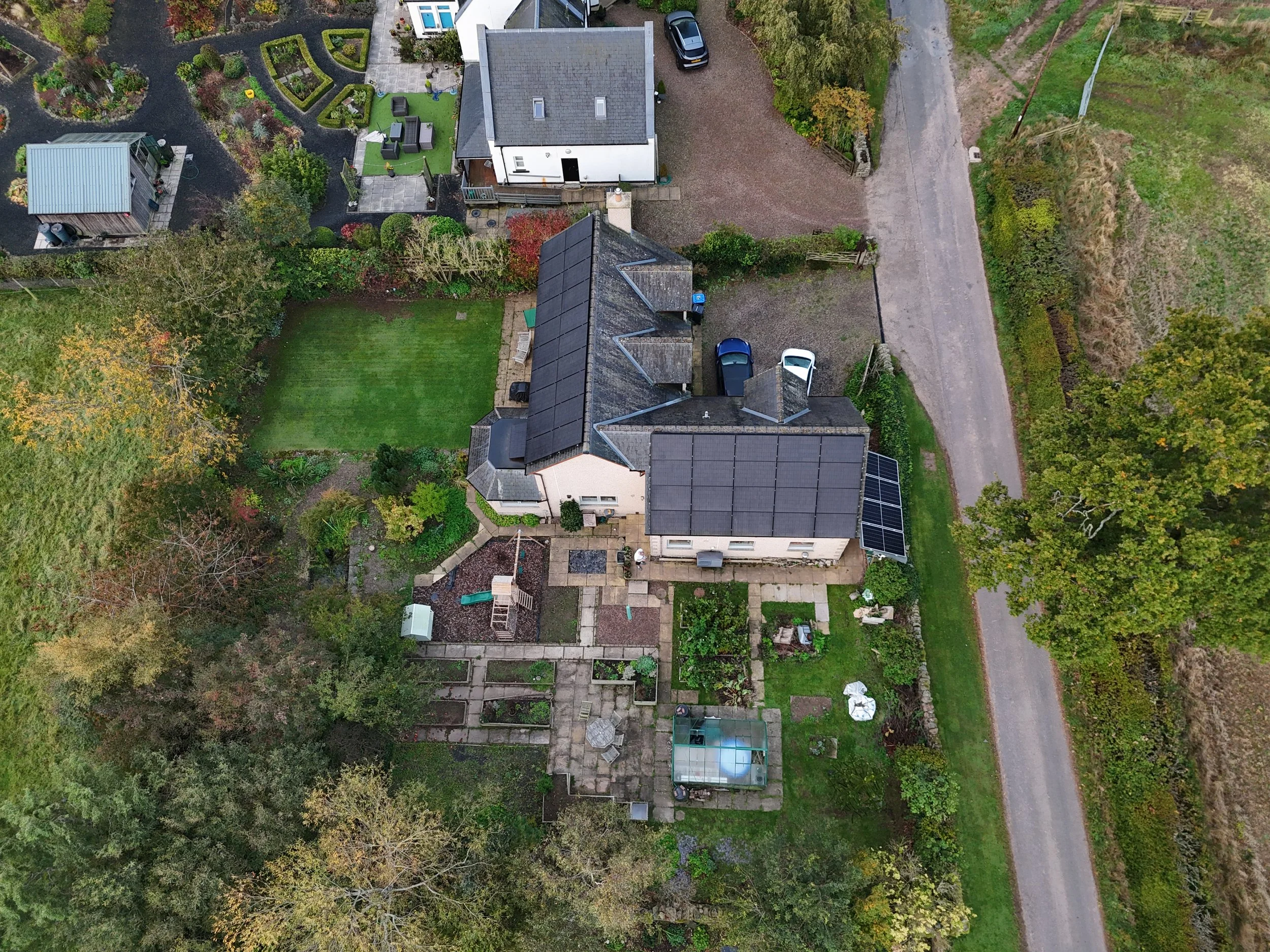 An aerial view of a residential backyard featuring a green lawn, a garden with flower beds, a patio area with outdoor furniture, a small shed, and several parked cars. A gravel driveway runs along the edge of the property, bordered by trees and veget