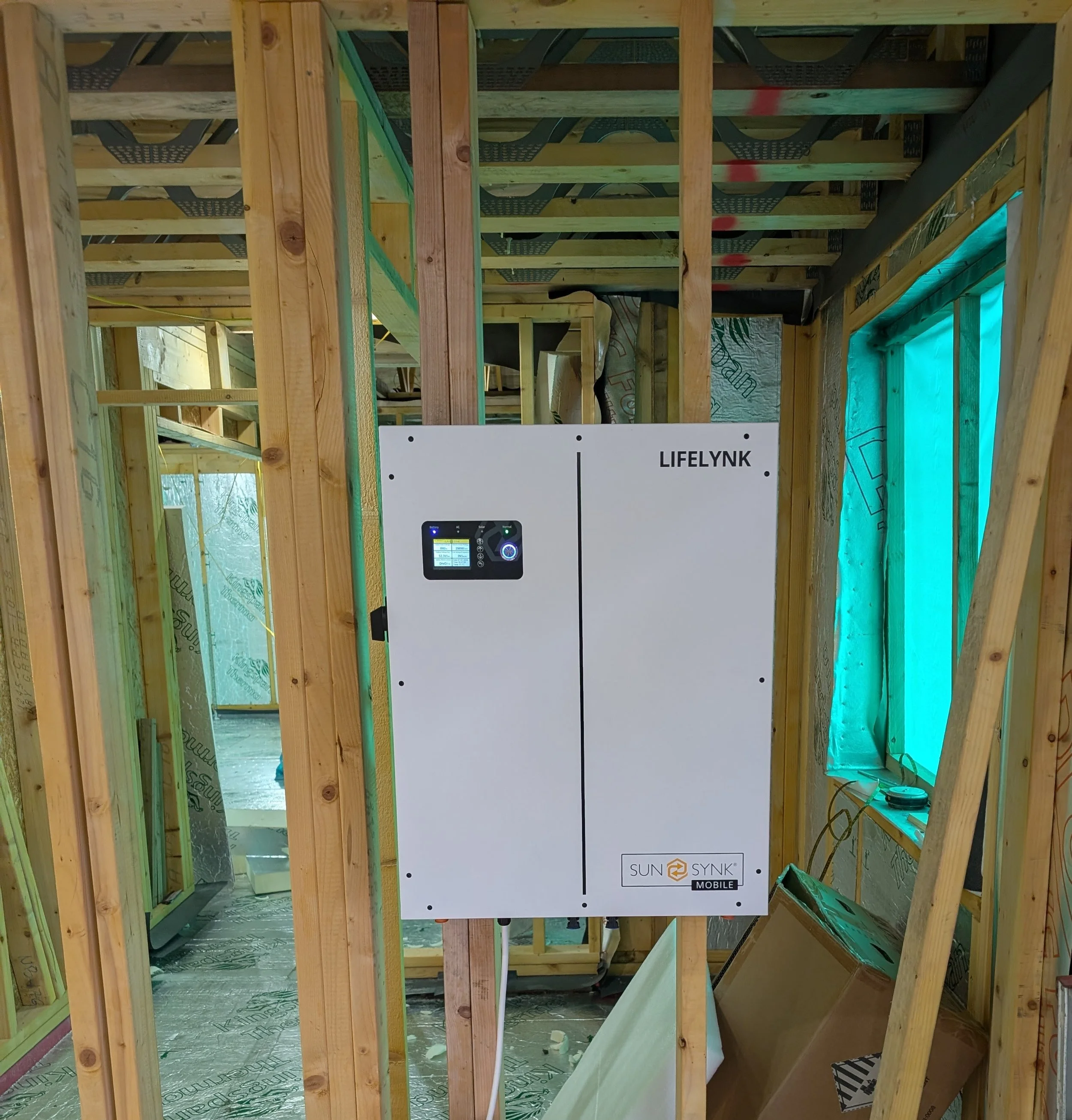 Construction framing with a white SunSynk mobile solar inverter mounted on the wall, in a building under construction with exposed wooden studs and green insulation material.