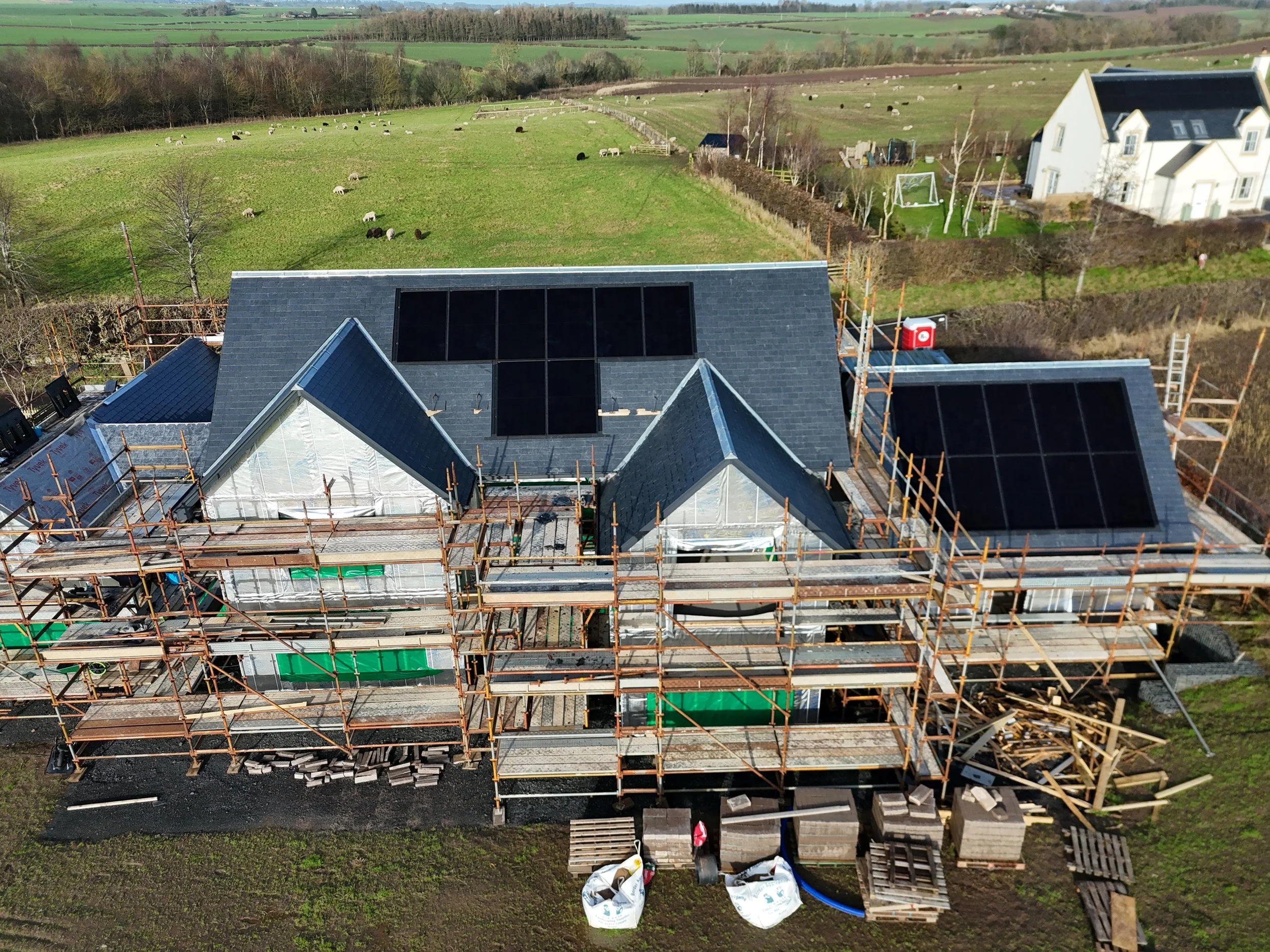 Aerial view of a house under construction with solar panels on the roof, surrounded by scaffolding, in a rural area with green fields and grazing sheep.