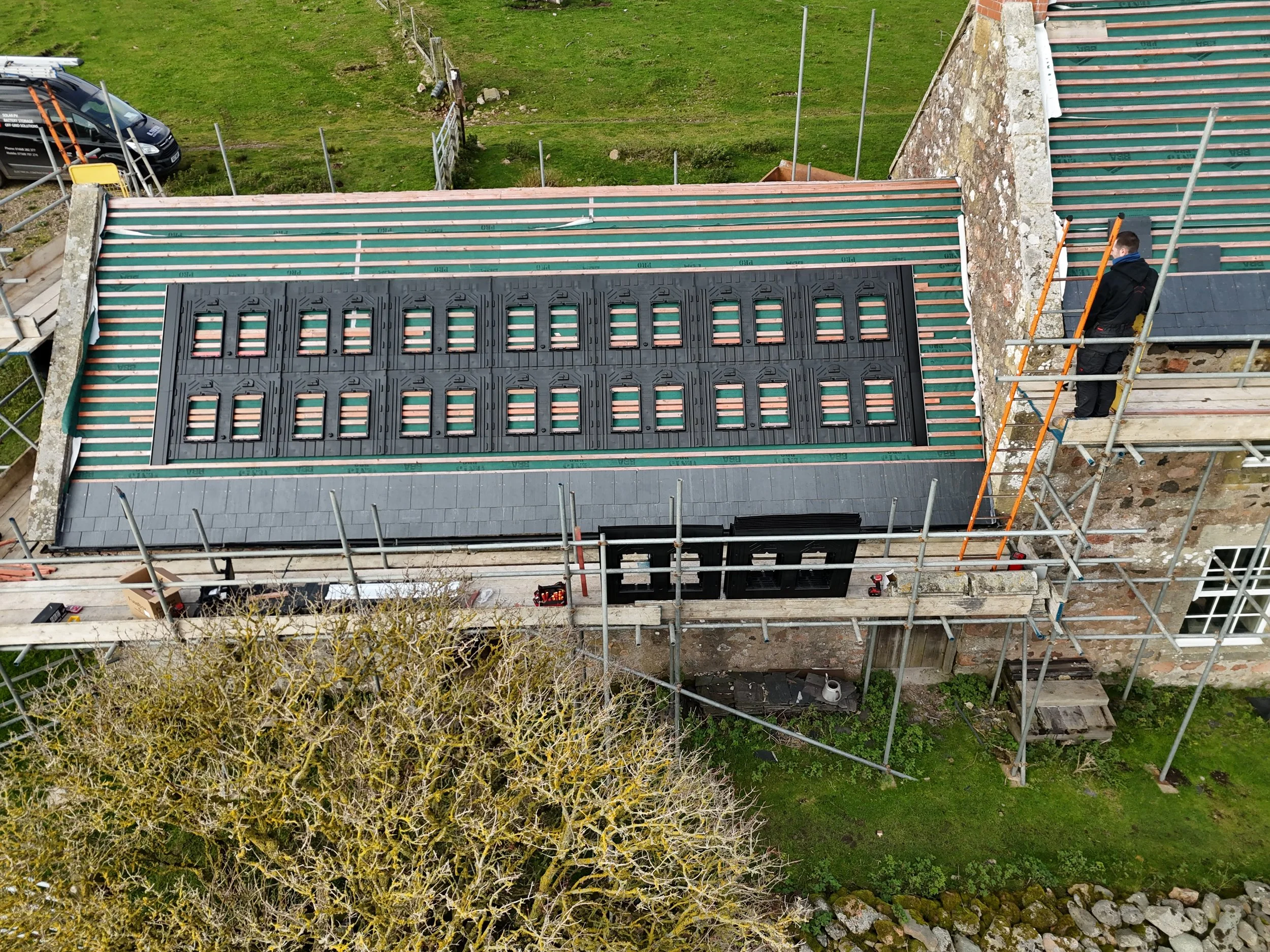 Aerial view of a roofing project showing a roof with new roofing materials and a worker in dark clothing working on the edge of the roof, with green grass and a parked car nearby.
