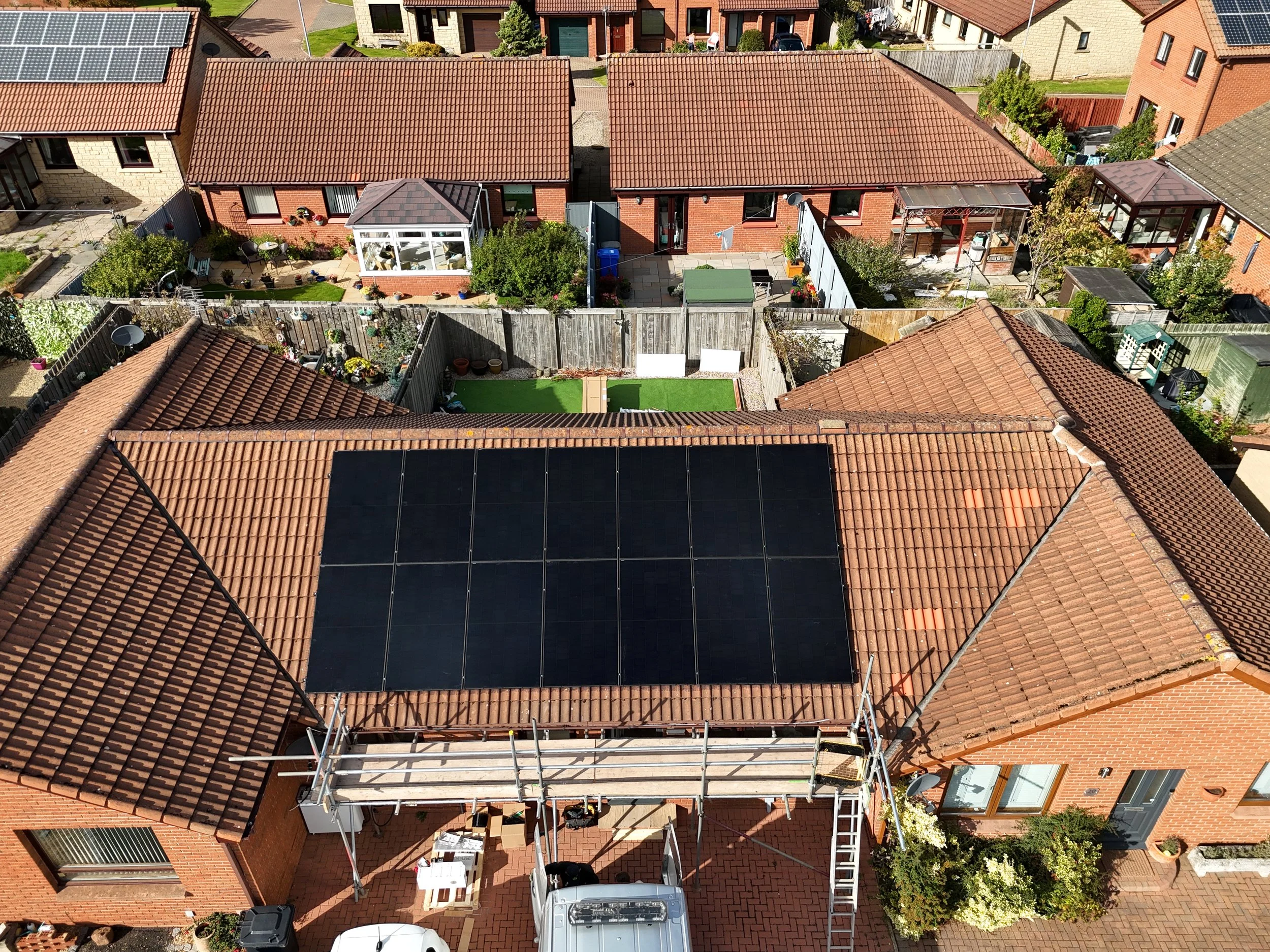 Aerial view of a row of houses with red brick walls and tiled roofs, some with solar panels, and a backyard garden with outdoor furniture, plants, and a small lawn.