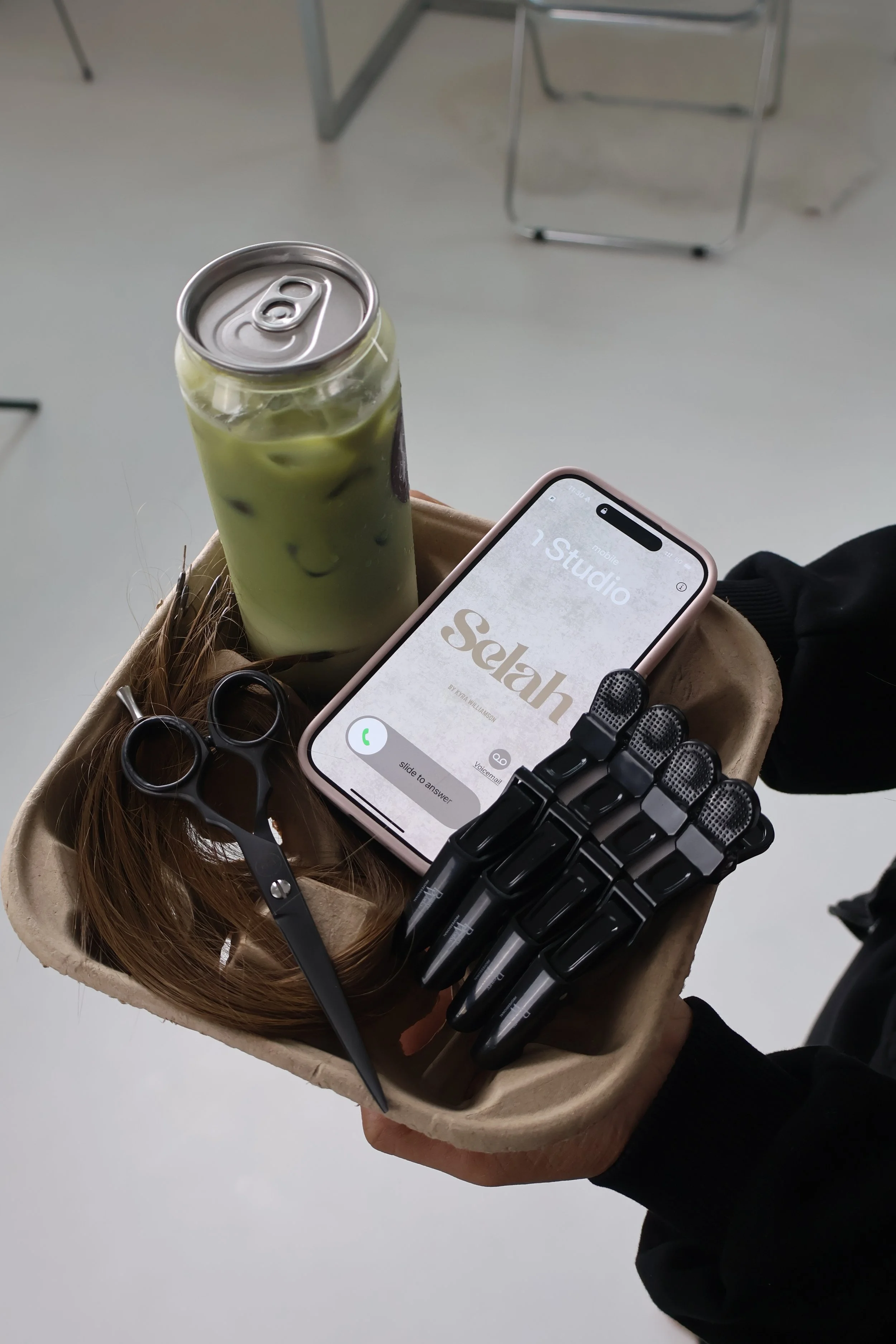 A disposable tray holding a can of iced matcha latte, a pair of black scissors, several black pens, and a smartphone displaying the word 'Selah' on its screen, all held by a person with black clothing.