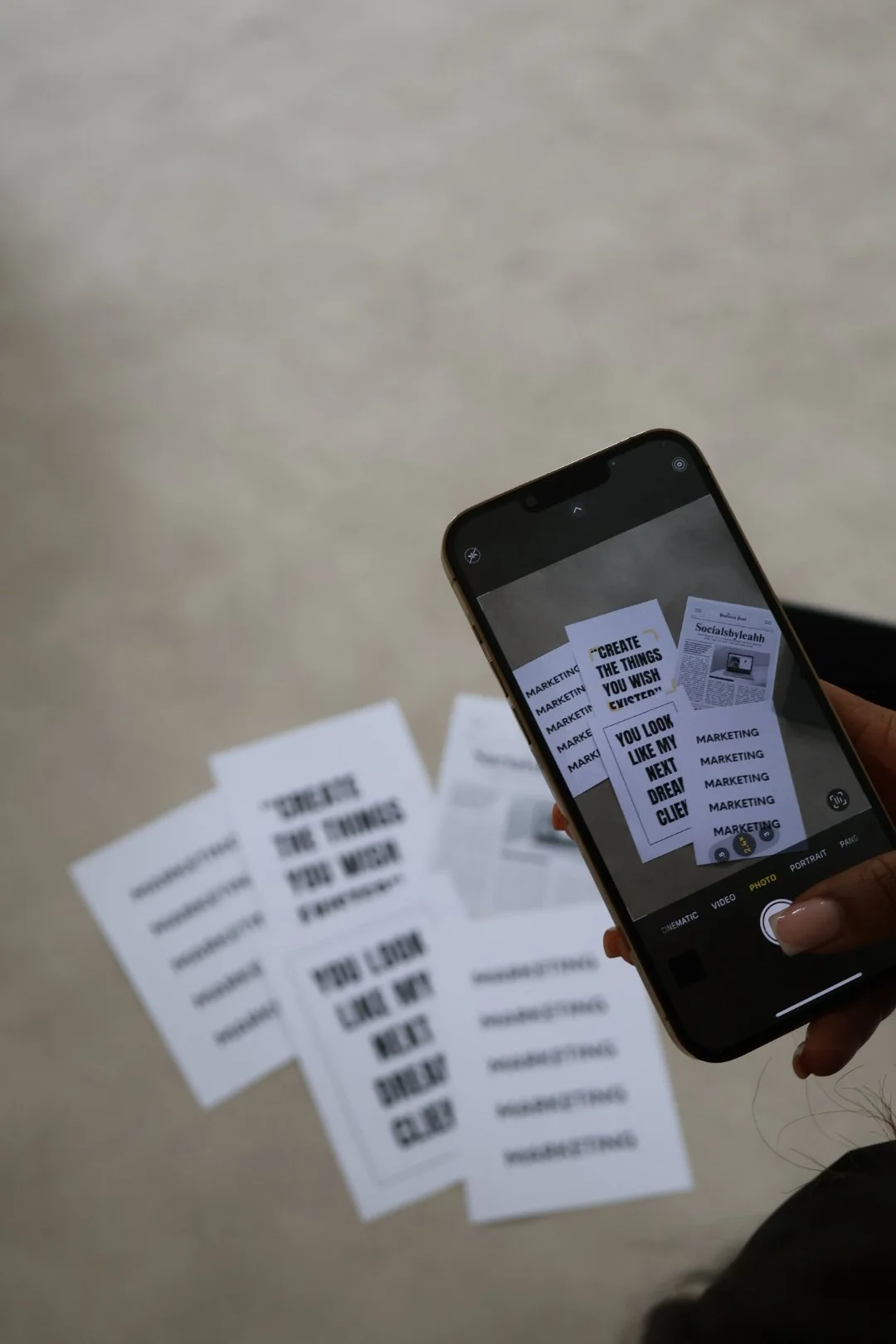 Person taking a photo of printed motivational and marketing quotes on paper sheets using a smartphone, with papers spread out on a beige surface.