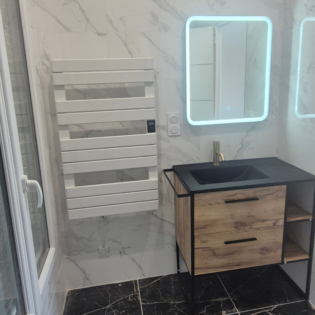 Salle de bain moderne avec miroir éclairé, lavabo noir en pierre, meuble en bois clair avec tiroirs, radiateur électrique blanc, et murs et sols en carrelage blanc marbré.