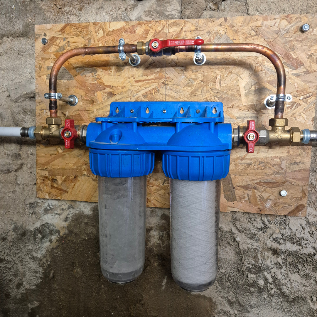Système de filtration d'eau domestique avec deux filtres, une plomberie en cuivre, et des valves rouges, fixé sur un panneau en bois contre un mur en pierre.