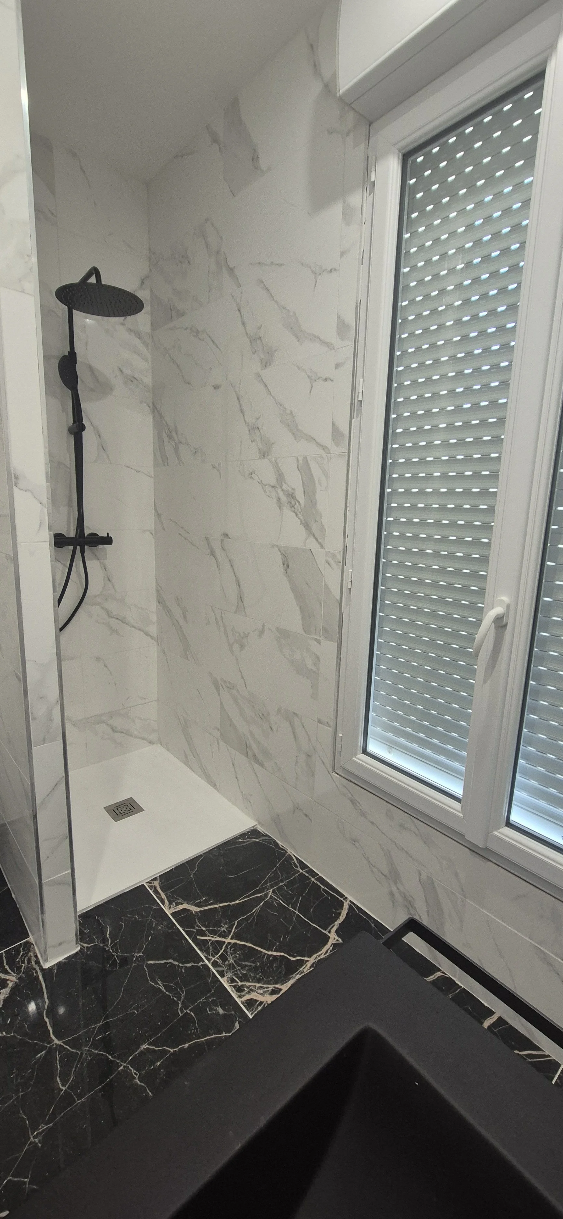 Salle de douche avec murs en marbre blanc et sol en marbre noir avec veines blanches. Douche avec pomme de douche noire et parois en verre partielle. Fenêtre avec volets roulants ferme.