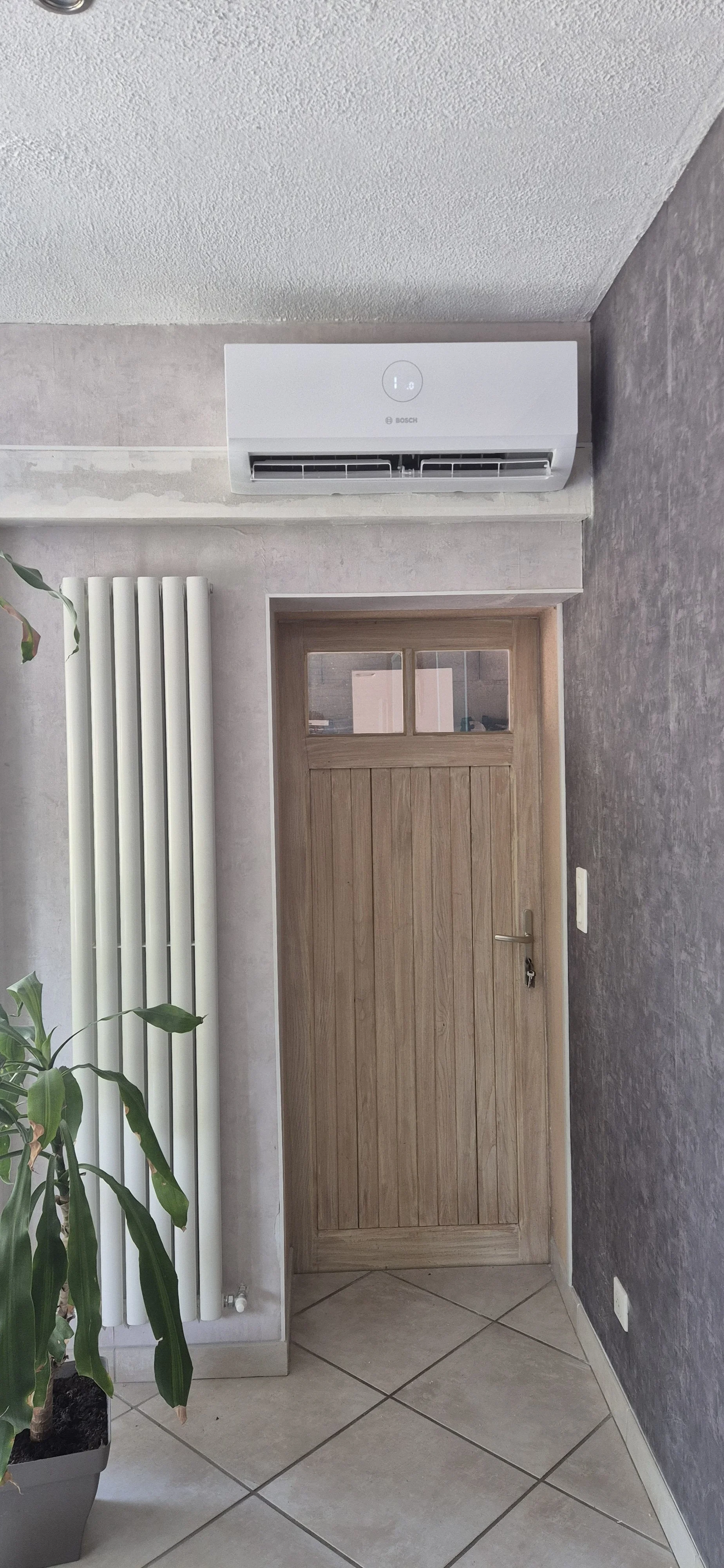 Une pièce intérieure avec une porte en bois, un radiateur blanc vertical, un climatiseur mural blanc Bosch et un pot de plante verte sur le sol en carreaux.