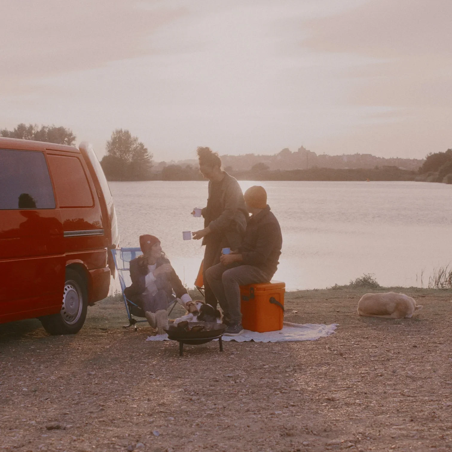 Family enjoying a picnic by a lake at sunset, with a red van parked nearby, a dog lying on the ground, and trees in the distance.