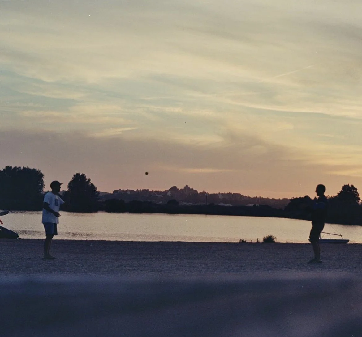 Two children playing with a ball by a lake at sunset, silhouette against a colorful sky.