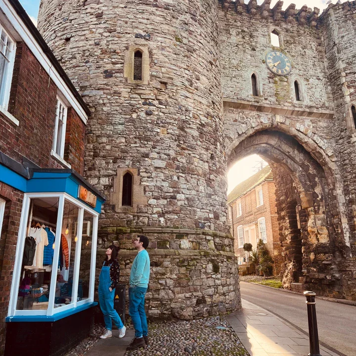 A historic stone city gate with an arched opening and a clock above it, with a shop window on the left and two people standing nearby.