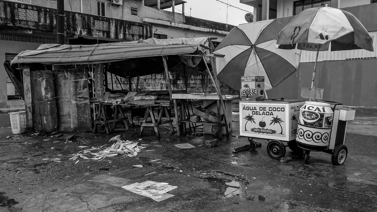 A small street-side stand selling coconut water and açaí, with umbrellas, signage, and some scattered trash on a wet pavement.