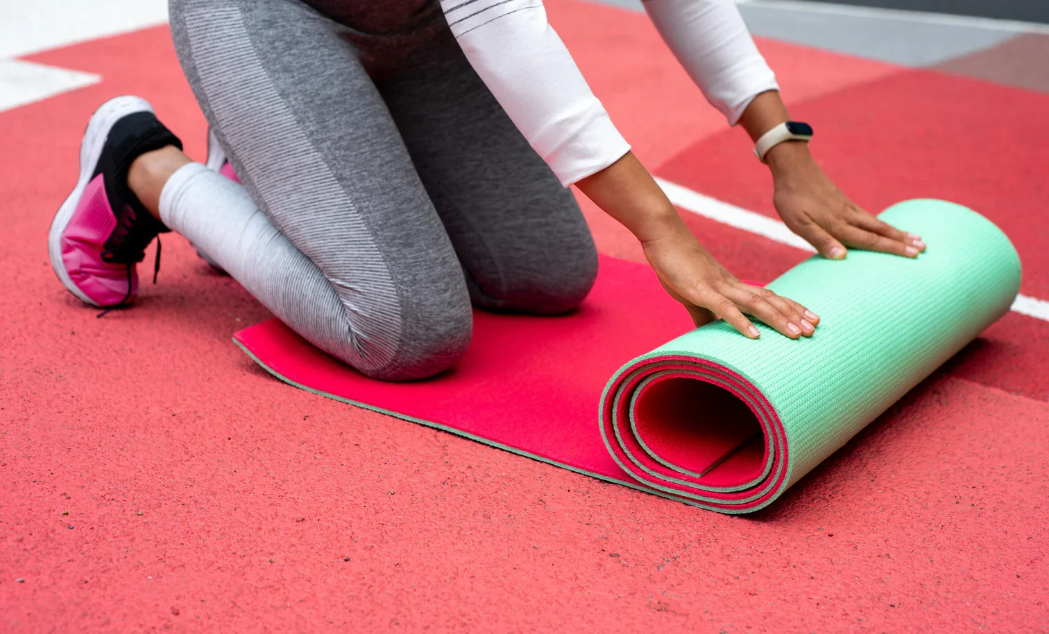 Person kneeling on a red running track, unrolling a green and pink yoga mat.