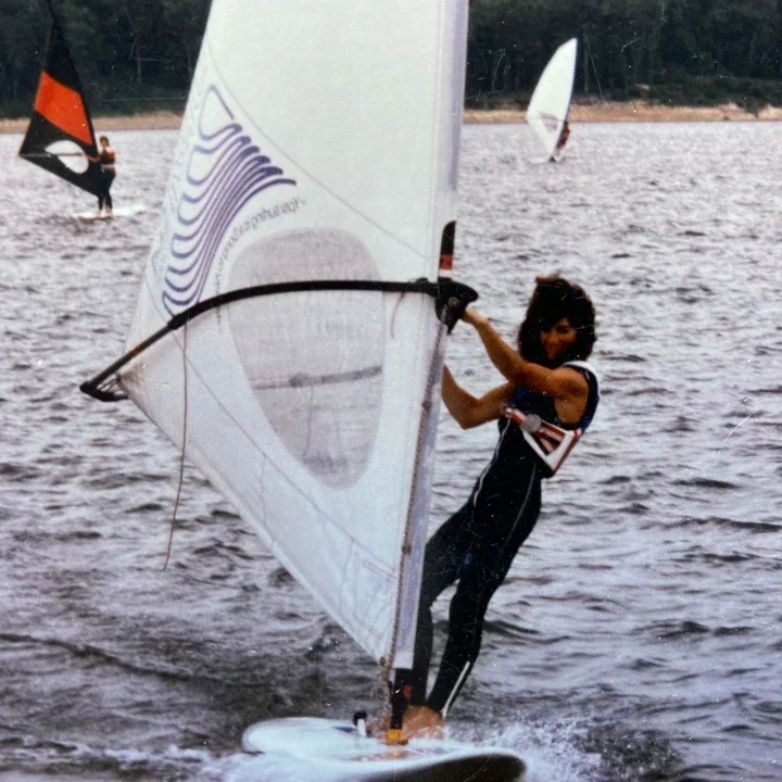 A woman windsurfing on a lake, holding onto the sail with a smile, with other windsurfers visible in the background.