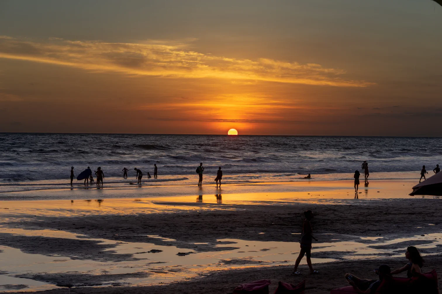 People enjoying the sunset on the beach, some walking, others playing in the water, with the sky painted in orange and yellow hues.