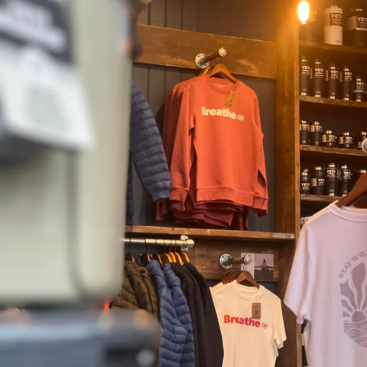 Pink sweatshirt with the word 'Breathe' hangs on a wooden rack in a store, with other jackets and T-shirts around.