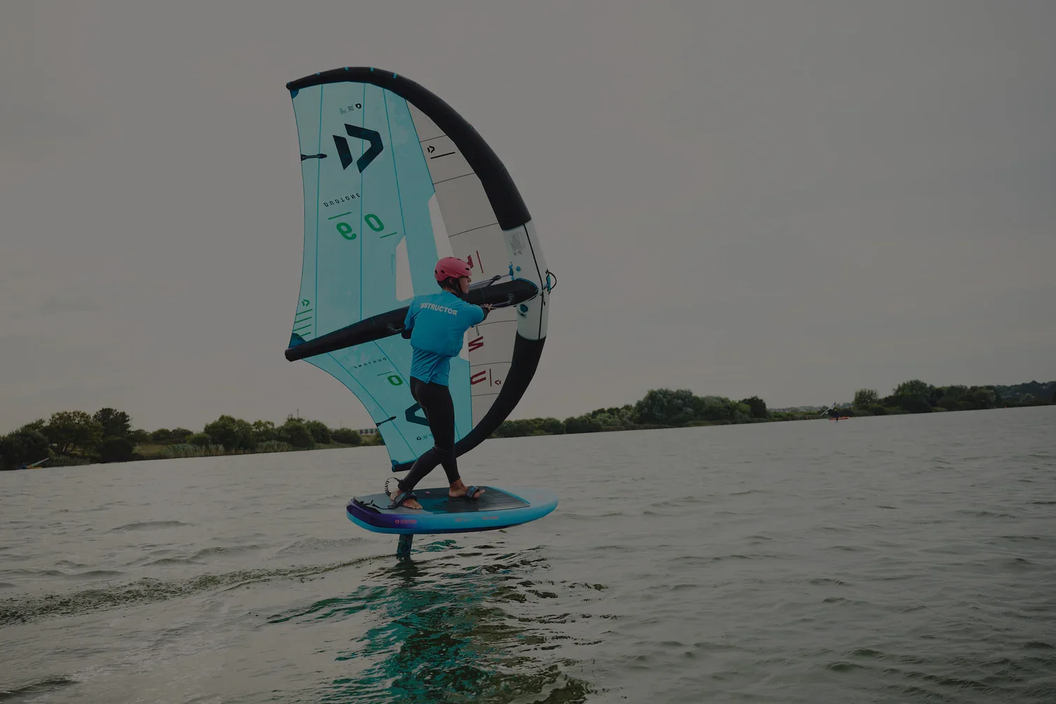 A person wearing a pink helmet and blue shirt riding a flyboard with an inflatable wing sail over a body of water on a cloudy day.
