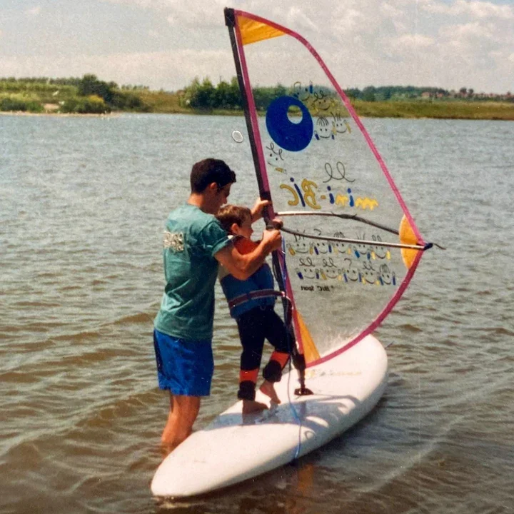 Two children learning to sail on a small windsurfing board in a lake, with an adult assisting them. The boy is holding the sail, and the girl is standing on the board. The scene is outdoors on a partly cloudy day.