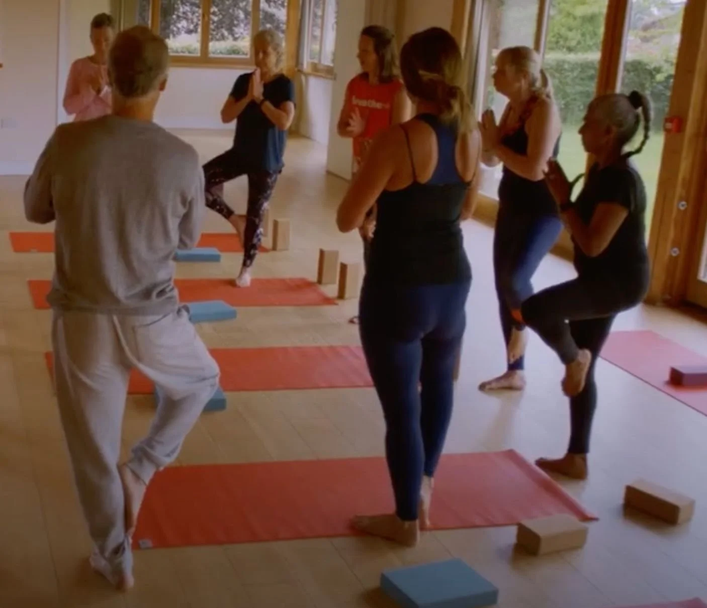 A group of women and a man practicing yoga indoors, balancing on one foot with hands in prayer position, on mats with bricks for support.