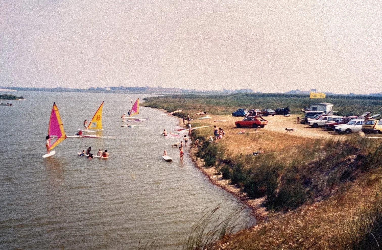 People enjoying a day at a lakeside, some sailing with colorful sails, others swimming and relaxing on the shore, with parked cars and a small gathering area on the grassy hill.