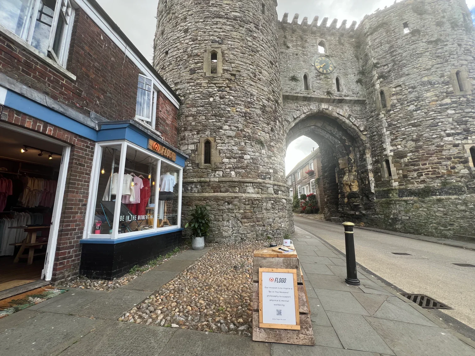 A street scene featuring an old stone castle gate with a clock, a retail store with clothing mannequins and a sign outside, and a small sidewalk cafe table with a note and QR code on a wooden stand.