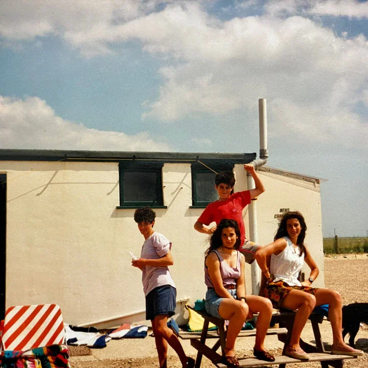 Four young people at the beach posing in front of a small building under a partly cloudy sky. Two girls are seated on a bench, one girl standing. One boy is standing holding a stick, another boy is leaning against the building.