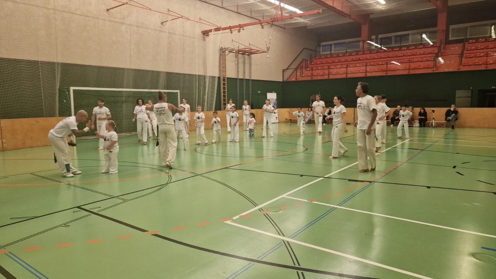 Menschen in weißen Kleidung beim Capoeira-Training in einer Turnhalle, Kinder und Erwachsene, einige sitzen an der Seite.