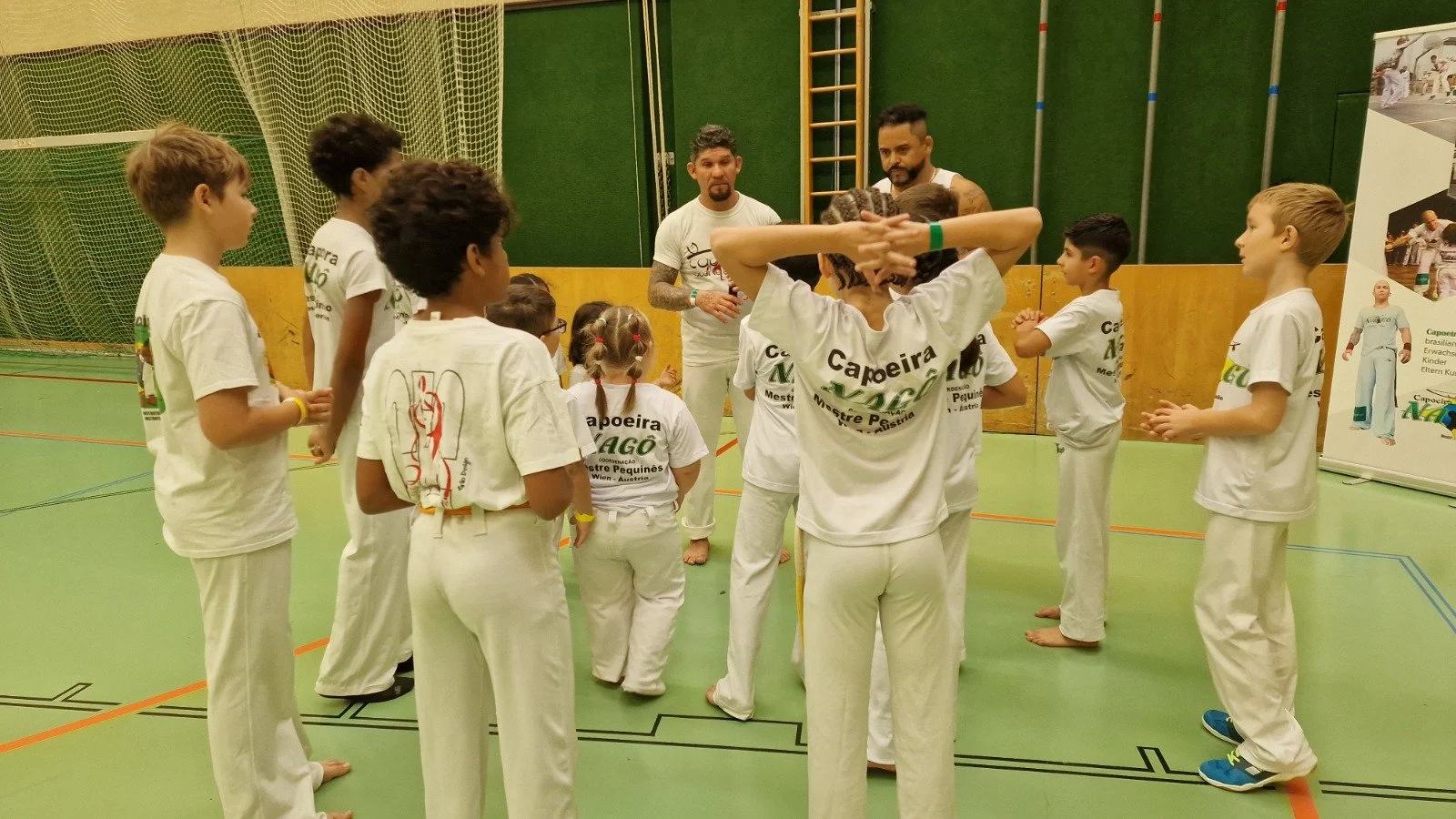 Gruppe von Kindern und Erwachsenen in einem Turnsaal, offensichtlich beim Capoeira-Training. Einige Kinder stehen in einem Kreis, während zwei Erwachsene, vermutlich Trainer, in der Mitte sprechen.