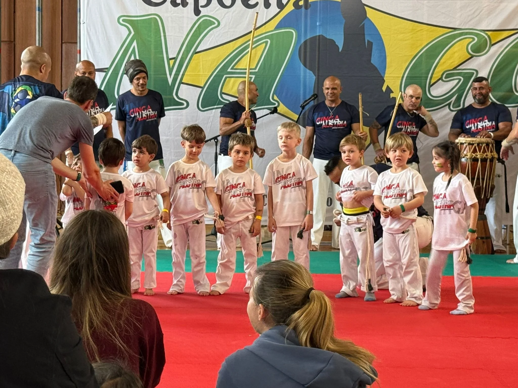 Kinder und Erwachsene beim Capoeira Event, Kinder stehen in Gürtelreihen, Erwachsene im Hintergrund mit Trommeln, große Werbefahne mit Nago Logo im Hintergrund.
