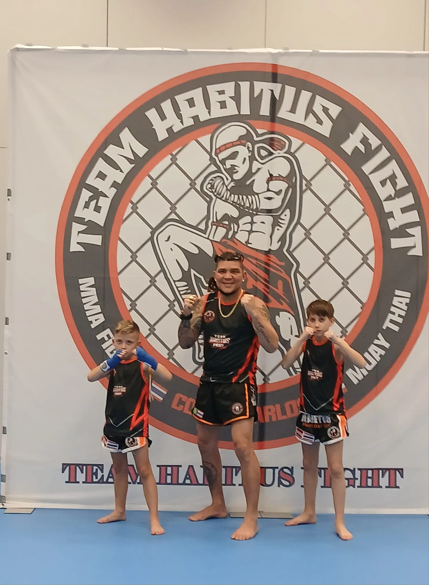 Mann und zwei Kinder im MMA-Kampfstil vor einem Logo mit einem Boxer in Kopfhaube, auf einem blauen Kampfboden.