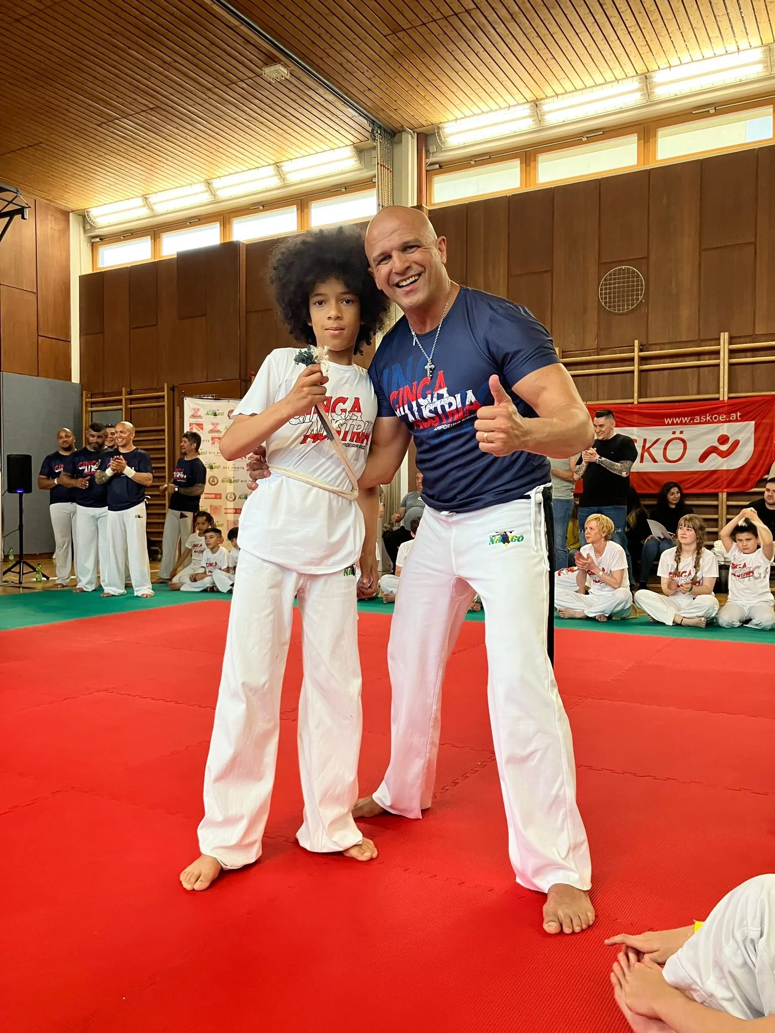 Zwei Menschen beim Capoeira in einem Turnraum, einer ist ein Junge mit Afro-Haaren und das andere ein Mann mit Glatze, beide tragen weiße Capoeira Kleidung, der Mann zeigt einen Daumen nach oben und lächelt, im Hintergrund sind weitere Kinder und Erwachsene, die Capoeira üben oder zuschauen.