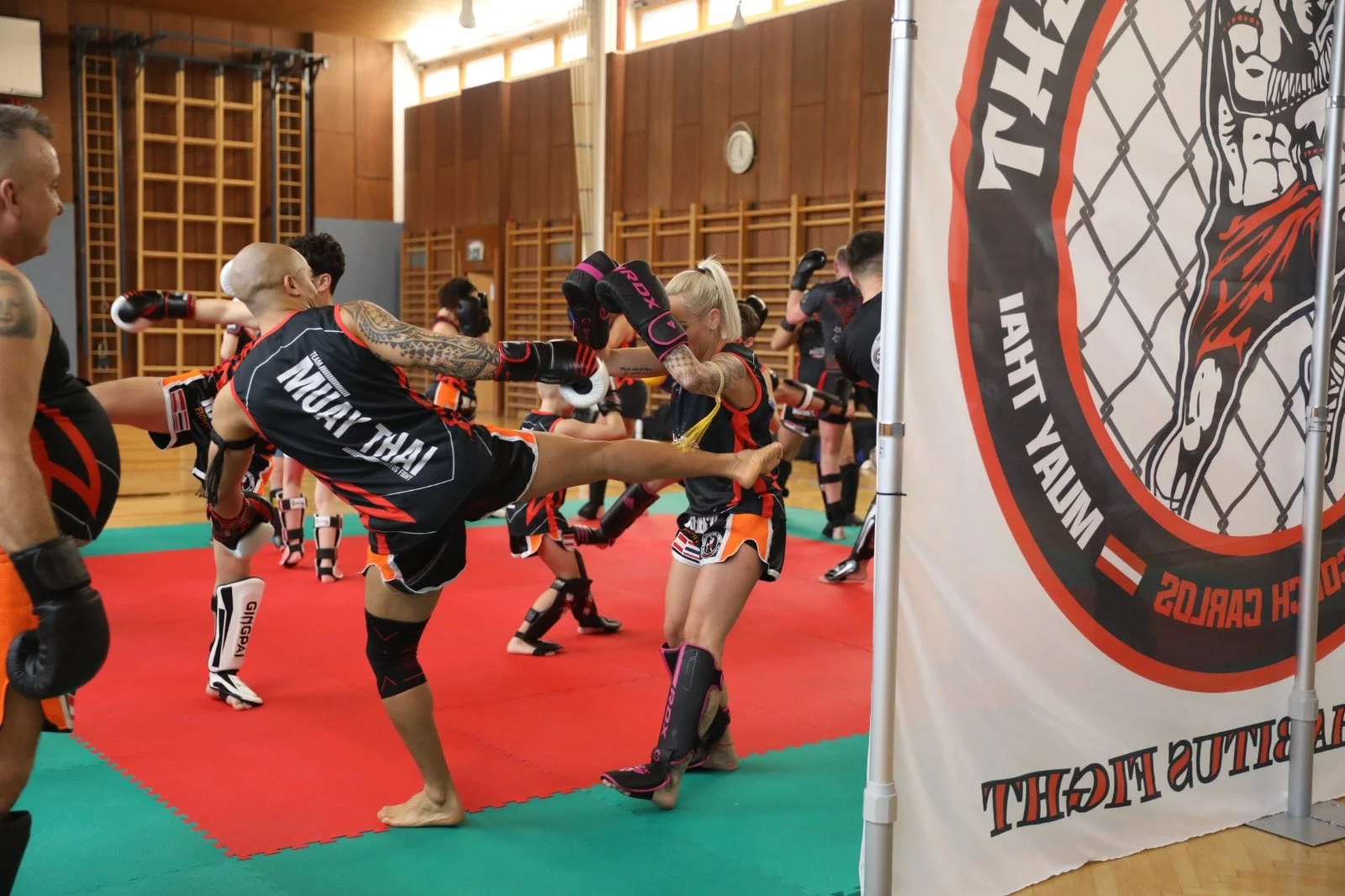 Muay Thai Kampfsporttraining mit mehreren Menschen in einem Dojo, die Techniken und Kicks üben, getragen in Muay Thai Ausrüstung, mit einem Banner an der Seite, das den Kampfsport zeigt.