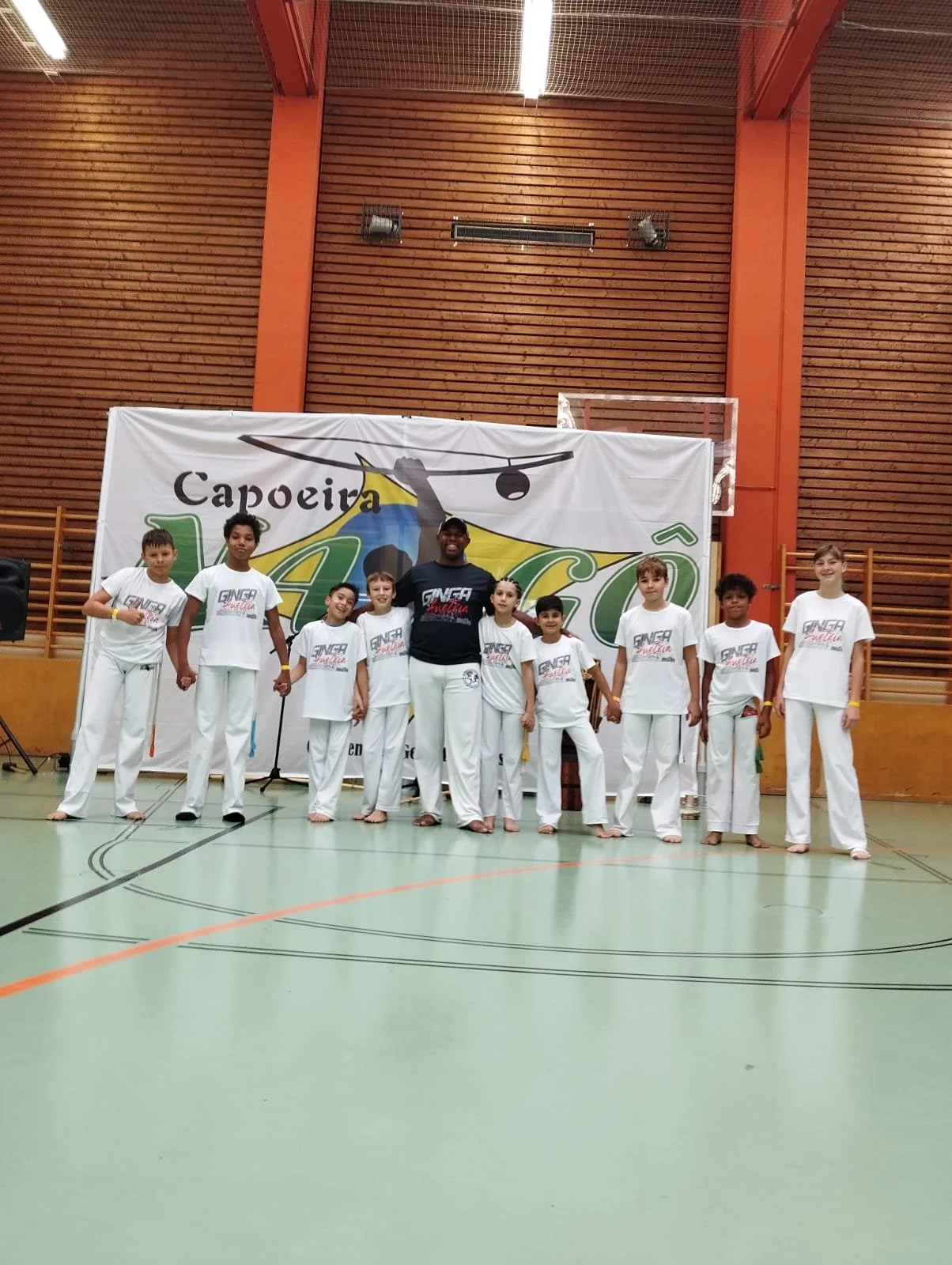 Gruppe von Kindern und einem Erwachsenen in weißen Capoeira-Uniformen, die in einer Turnhalle vor einem Banner stehen.