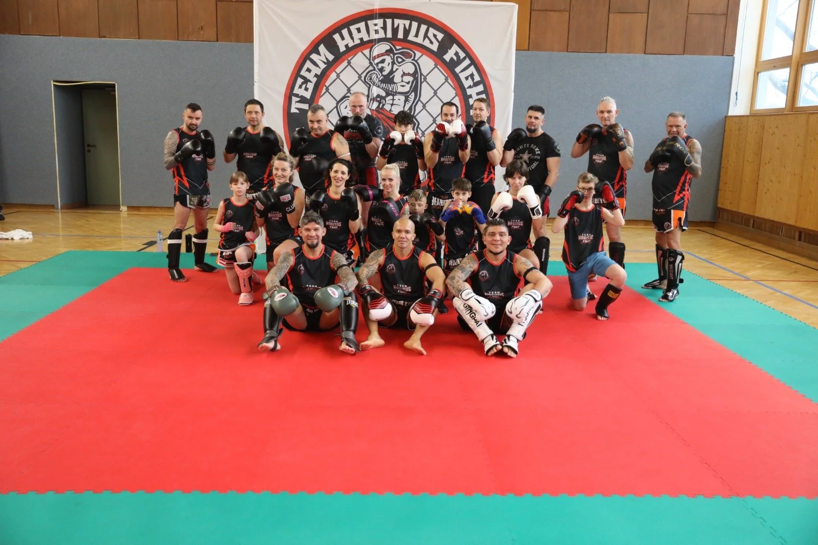 Gruppe von Kickboxern und Trainern in einem Gym, alle tragen Trainingskleidung mit schwarzen und roten Farben, die meisten tragen Handschuhe oder Schutzausrüstung, im Hintergrund ein Banner mit einem Kampfsport-Logo.