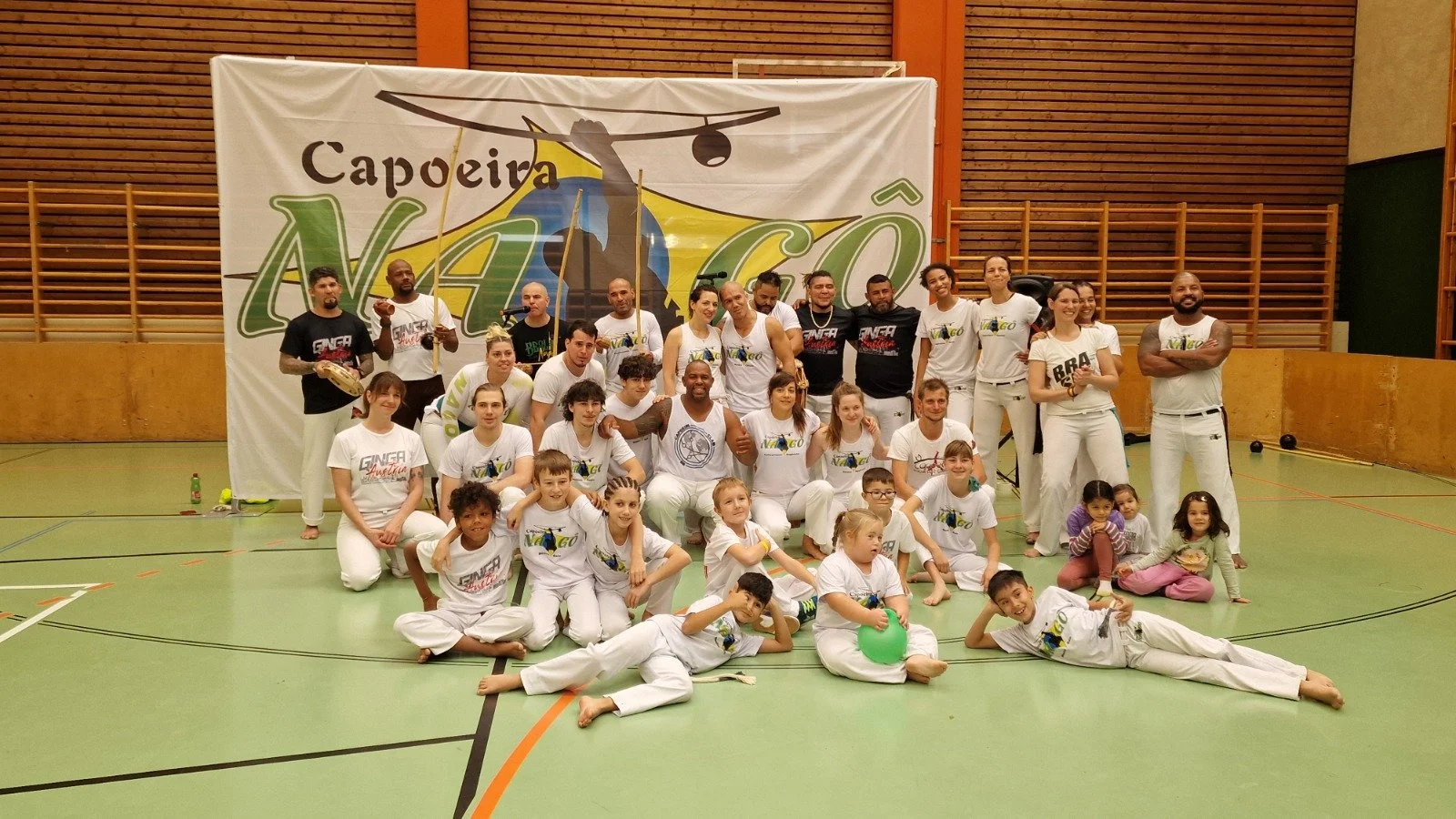 Gruppe von Menschen in weißen und schwarzen Capoeira-Kleidung in einer Turnhalle, vor einer großen Capoeira-Nach-Ó-Banner, die eine Capoeira-Figur und den Globus zeigt, beim Capoeira-Workshop oder -Event.