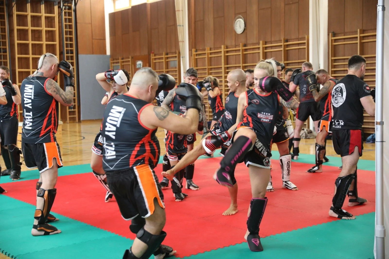 Trainingsszene einer Kampfsportgruppe in einer Turnhalle mit Holzvertäfelung, alle in schwarzen und roten Uniformen, einige in Schutzausrüstung bei Training auf weichem Untergrund.