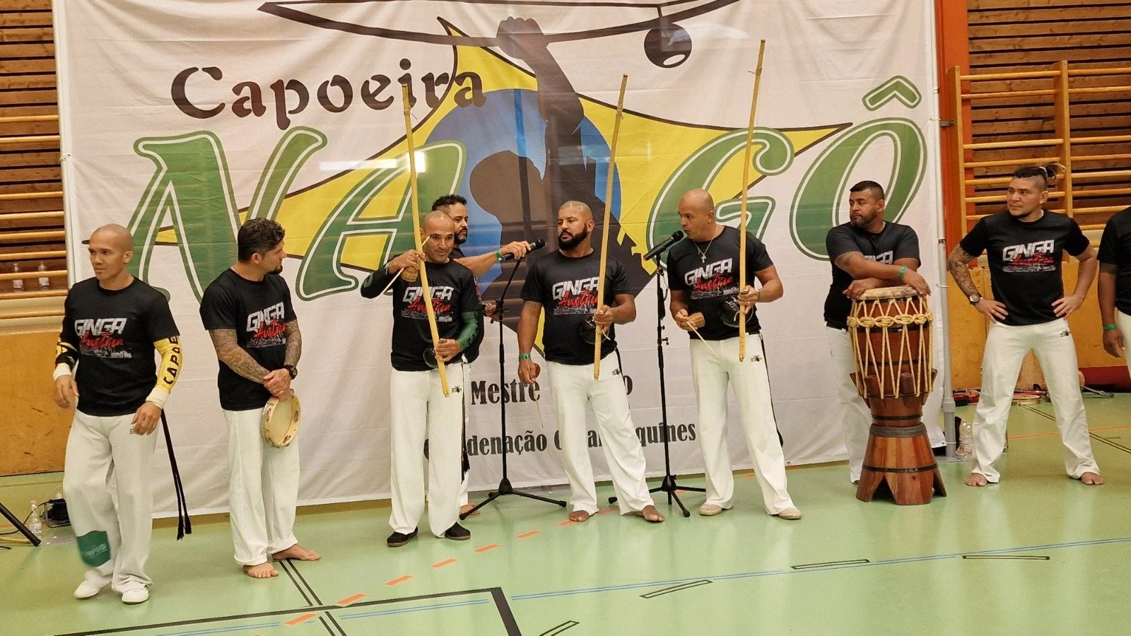 Gruppe von Capoeira-Kämpfern in schwarzen T-Shirts und weißen Hosen, einige halten Musikinstrumente und stehen vor einem Bühnenhintergrund mit dem Wort 'Capoeira' und 'Nação'.