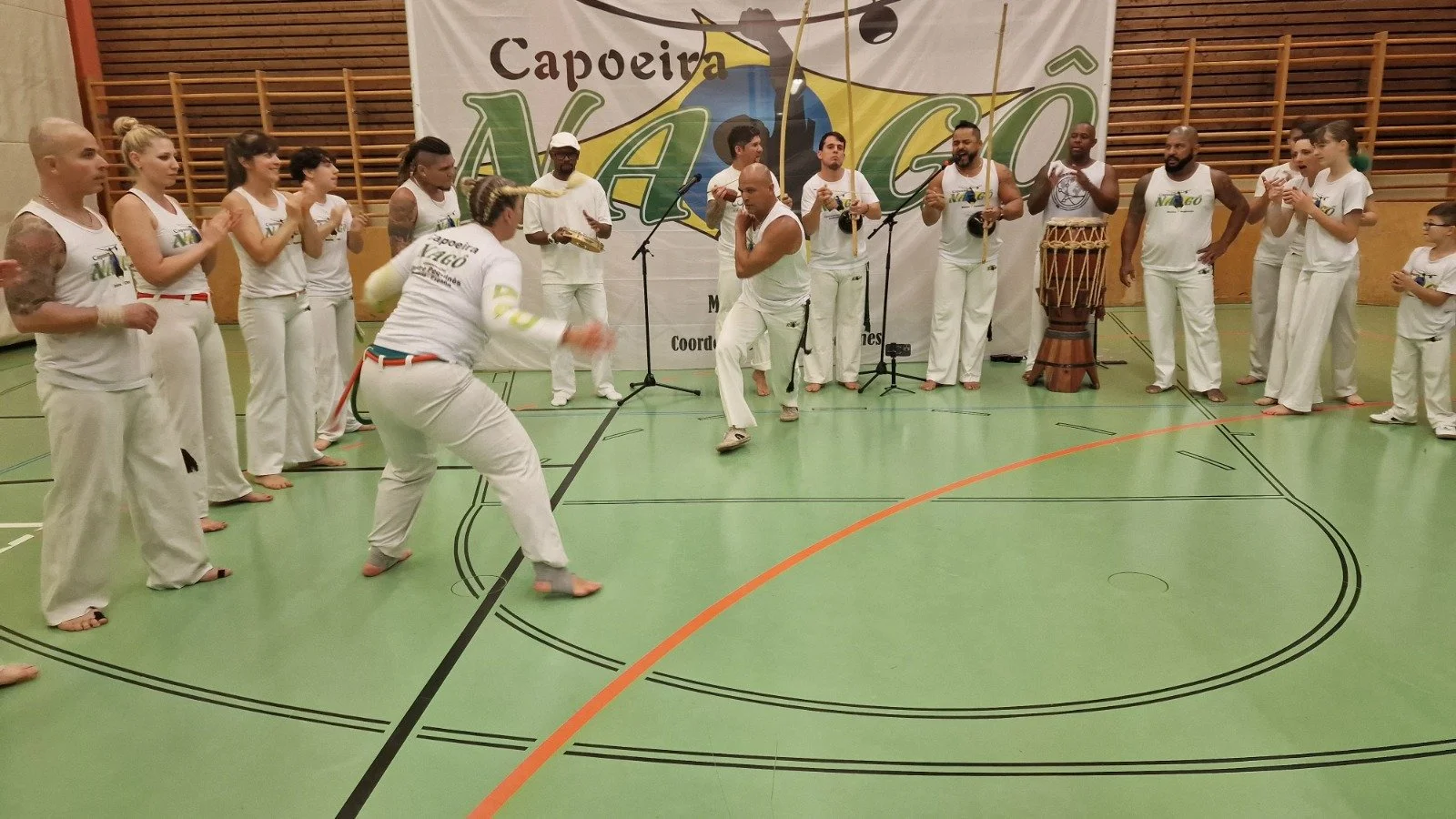 Gruppe von Menschen beim Capoeira-Training in einer Turnhalle, mit Musikern und Tänzern, die eine Capoeira-Aufführung machen.