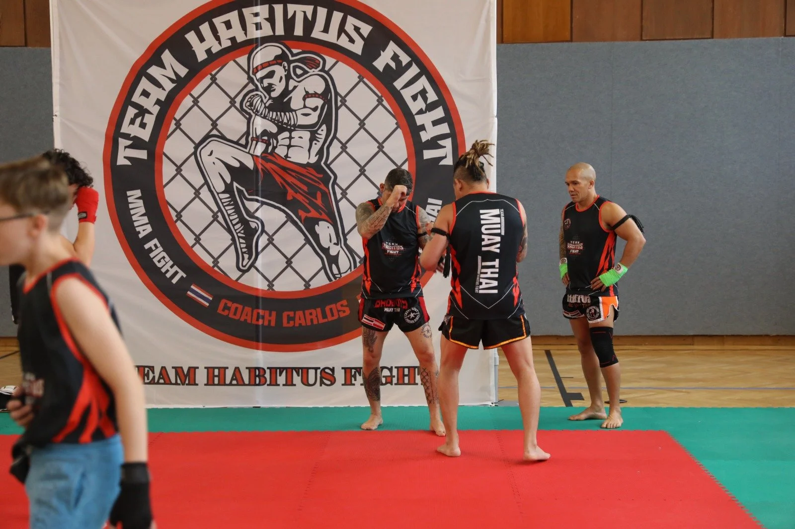 Muay Thai Kämpfer trainieren vor einem Banner mit dem Logo "Team Habitus Fight" und "Coach Carlos" in einer Turnhalle, während ein weiterer Kämpfer zuschaut.