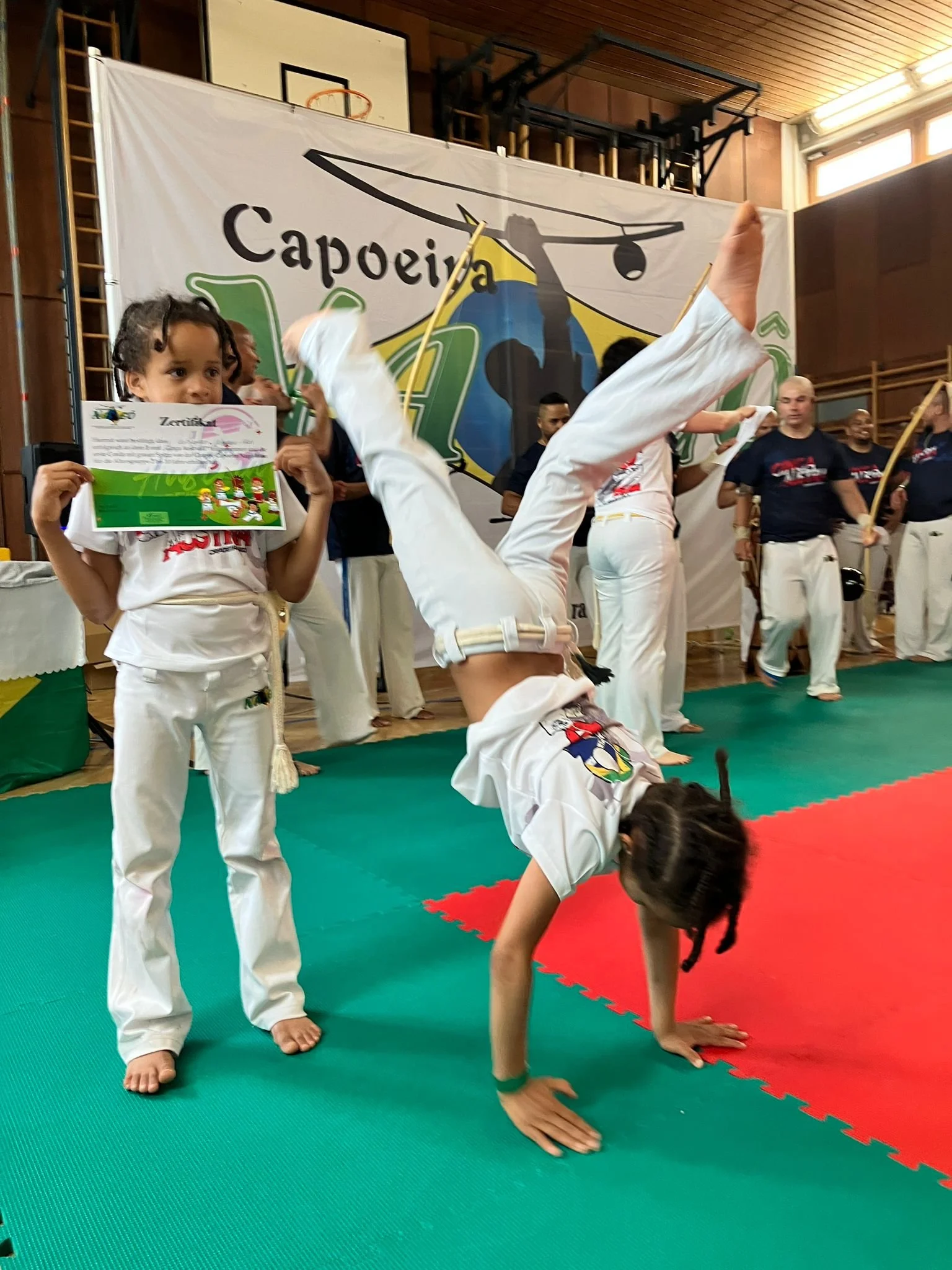 Eine Gruppe von Menschen in Karate-Uniformen übt in einer Turnhalle. Ein Kind macht einen Handstand auf der Matte, während andere im Hintergrund stehen. Ein Mädchen hält eine Urkunde in der Hand.